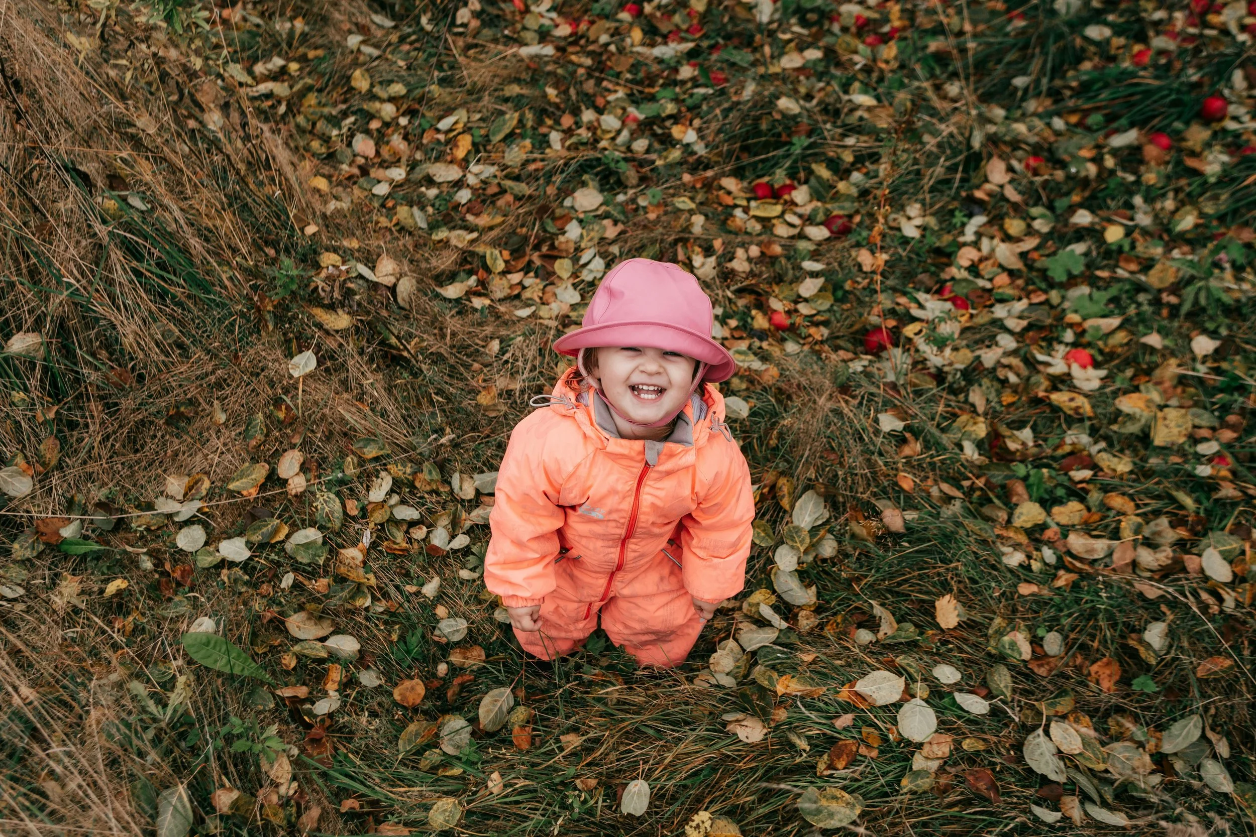 A young girl in an orange jacket and pink hat smiling and looking up at the camera, standing in autumn leaves.