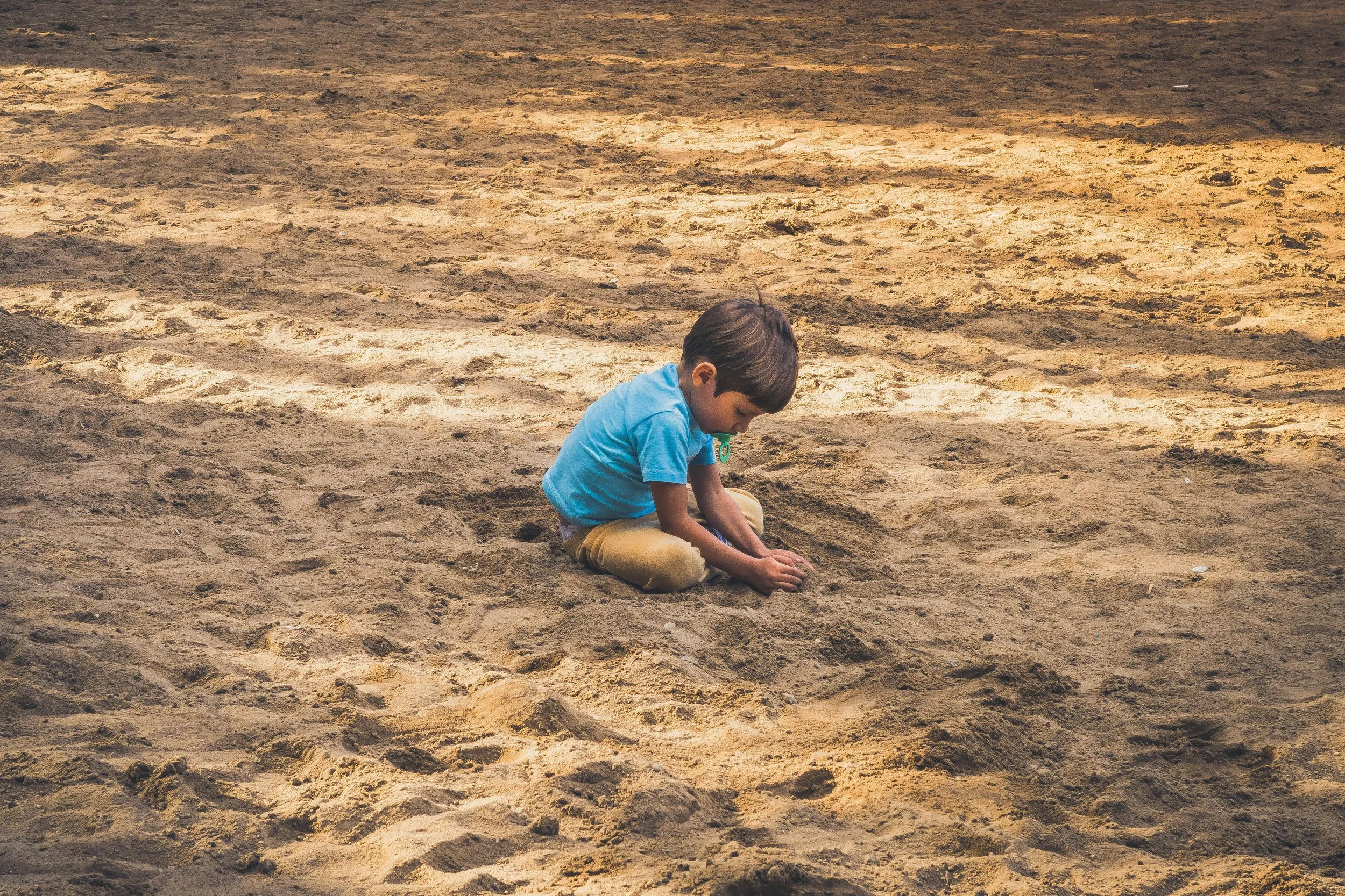 A young boy with brown hair, wearing a blue shirt and yellow pants, sits on sandy ground and plays with the sand using his hands.