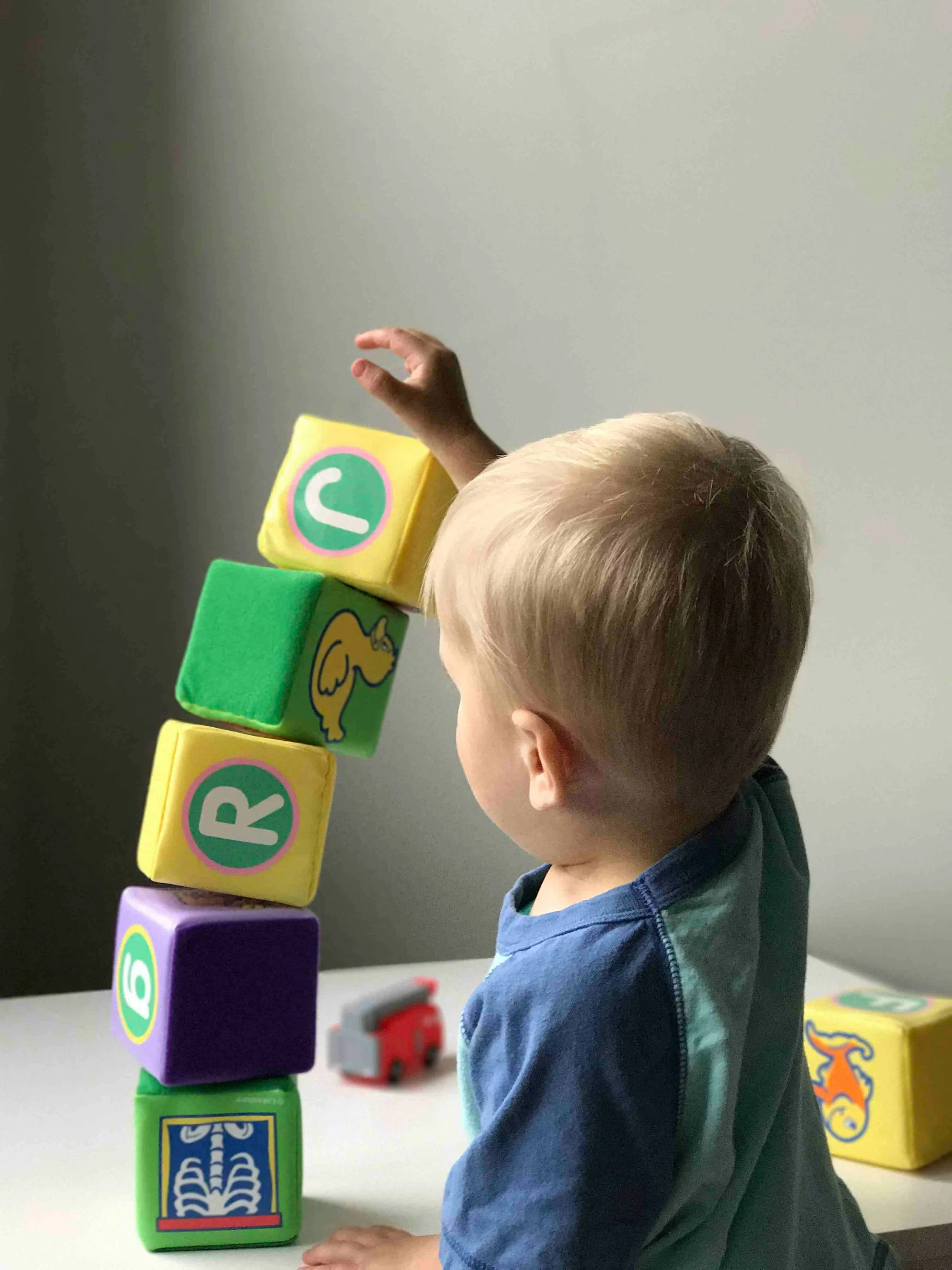 A young boy playing with colorful foam alphabet blocks stacked vertically, reaching with his right hand to add a block at the top. There are additional toys on the table, including a small red vehicle, and a yellow box with a dinosaur illustration. The background is a plain light-colored wall.