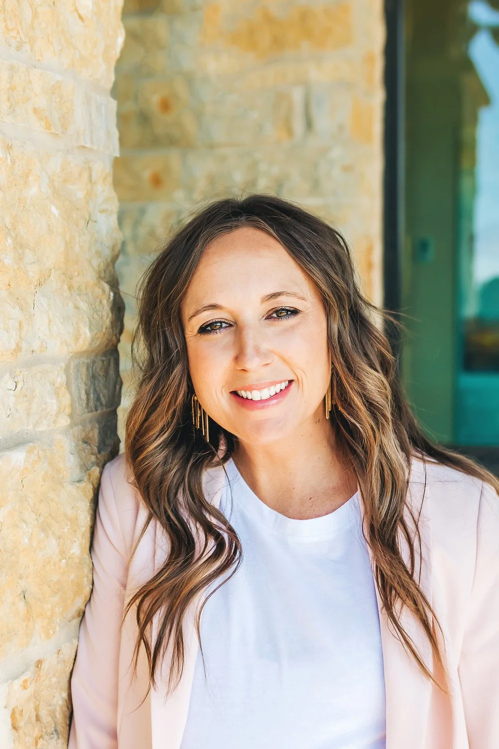 Smiling woman with wavy brown hair wearing earrings and a light pink blazer, leaning against a stone wall, outdoors with a reflective glass window behind her.