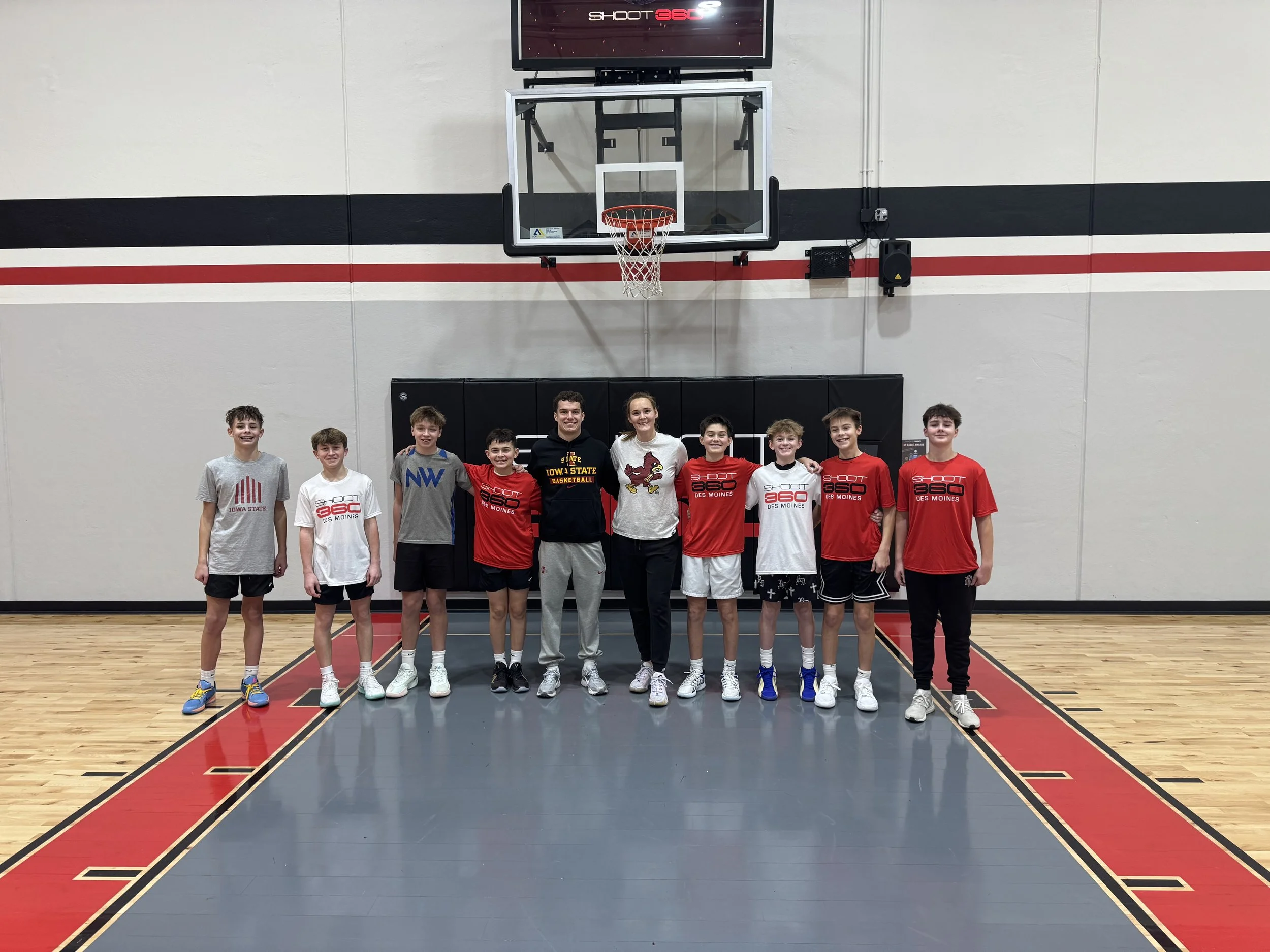 A group of ten young boys and girls standing in a basketball court, posing for a photo. They are dressed in athletic clothing, some with Iowa State and Des Moines basketball shirts, and are smiling at the camera in front of a basketball hoop and scoreboard.