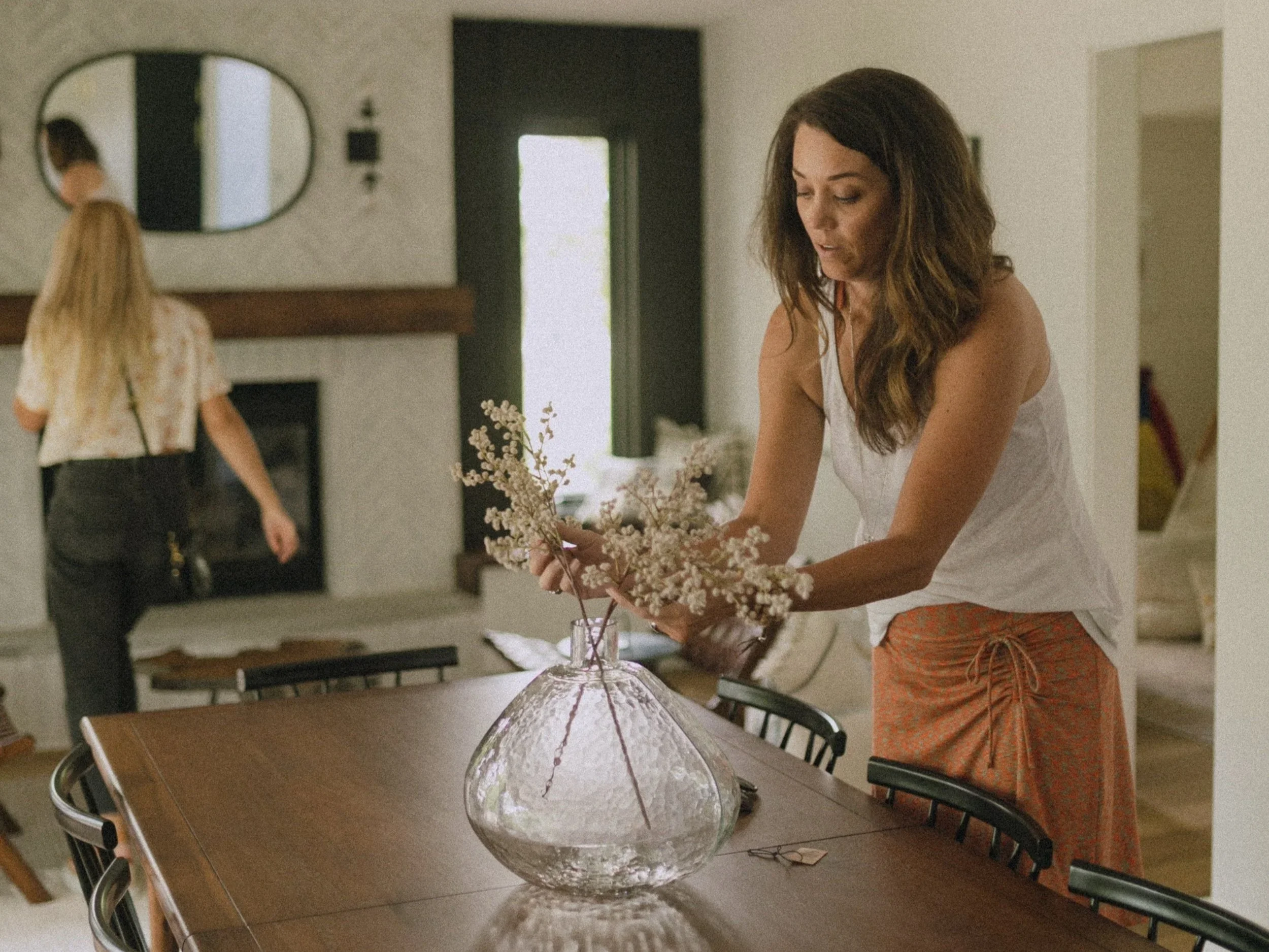 A woman with long dark hair arranging a flower arrangement in a large glass vase on a dining table, with another woman in the background near a fireplace and mirror.