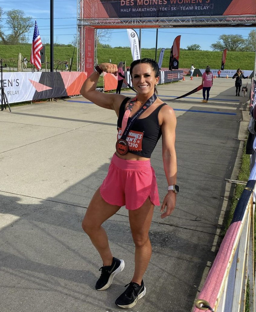 A smiling woman in athletic attire, wearing a black sports bra and pink running shorts, proudly displaying a medal at a race event. She is standing outdoors near the finish line of a race, with banners and flags in the background, and holding the medal to her neck.