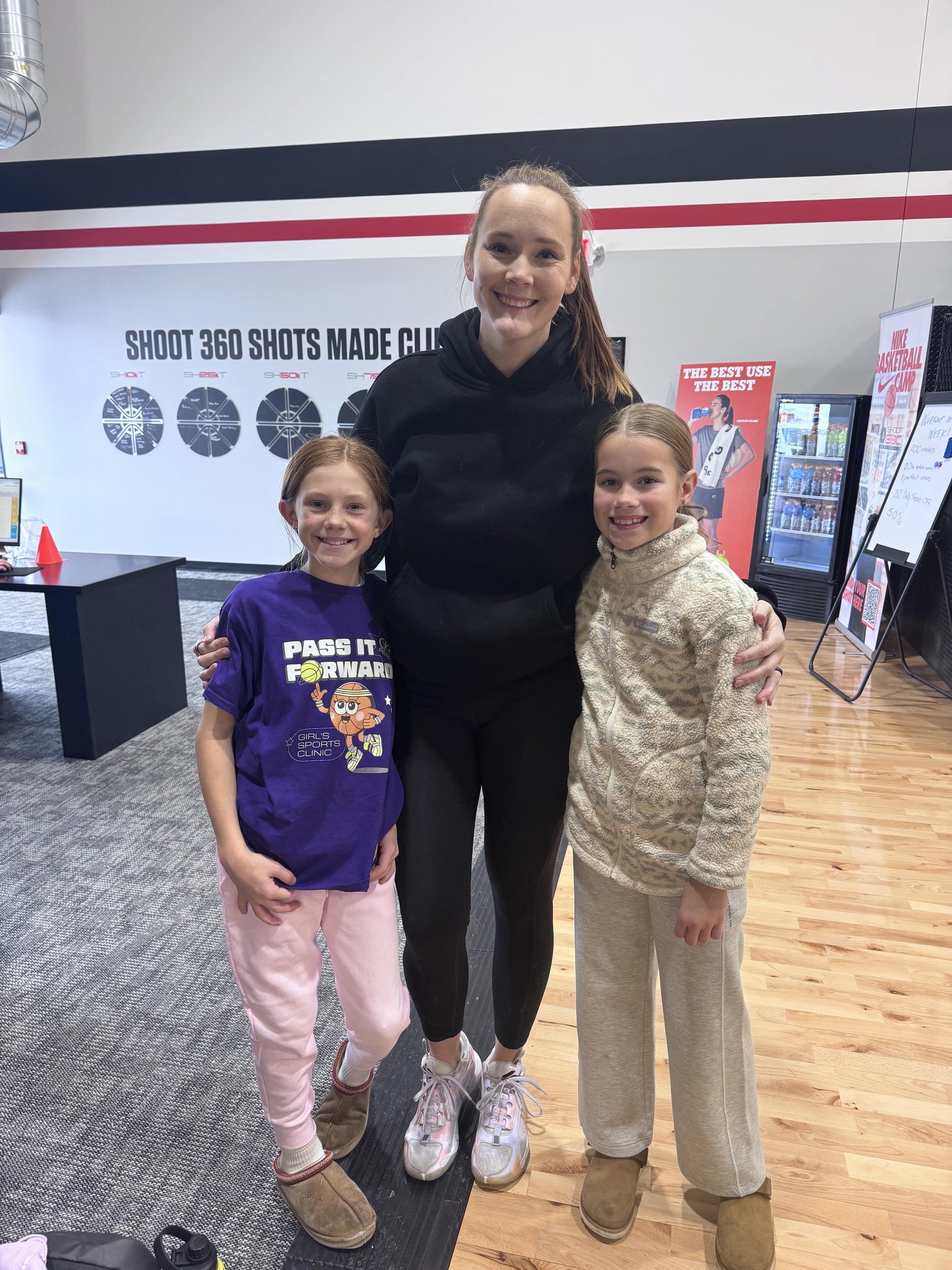 Three young girls and a woman standing together indoors in a fitness or sports facility, smiling at the camera. Two of the girls are on either side of the woman, and one girl is to her left. The background features signs and a vending machine.