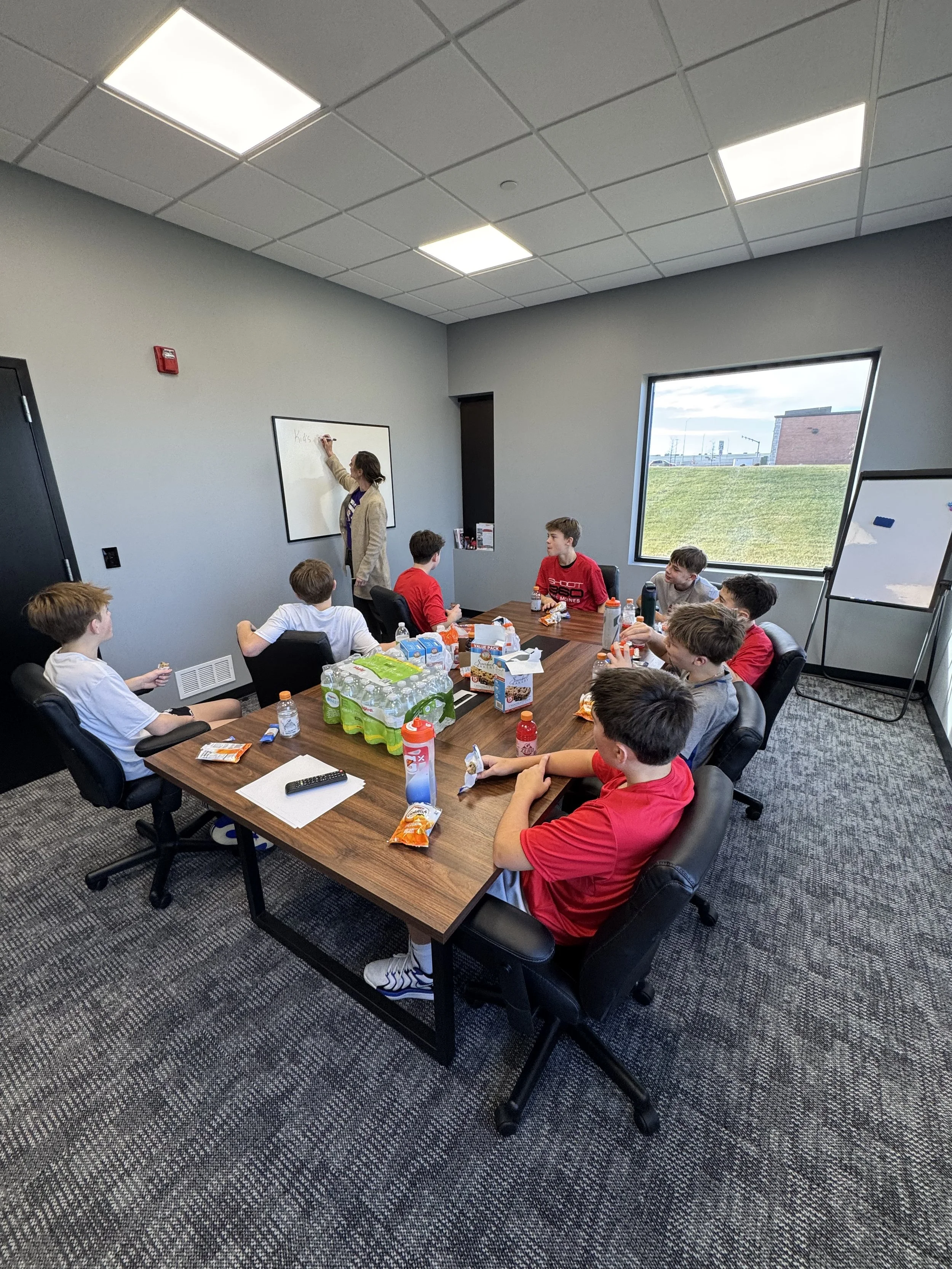 A classroom meeting with a teacher writing on a whiteboard and students seated around a table with snacks and drinks, in a room with gray walls, carpeted floor, a window showing an outdoor scene, and a whiteboard on a stand.