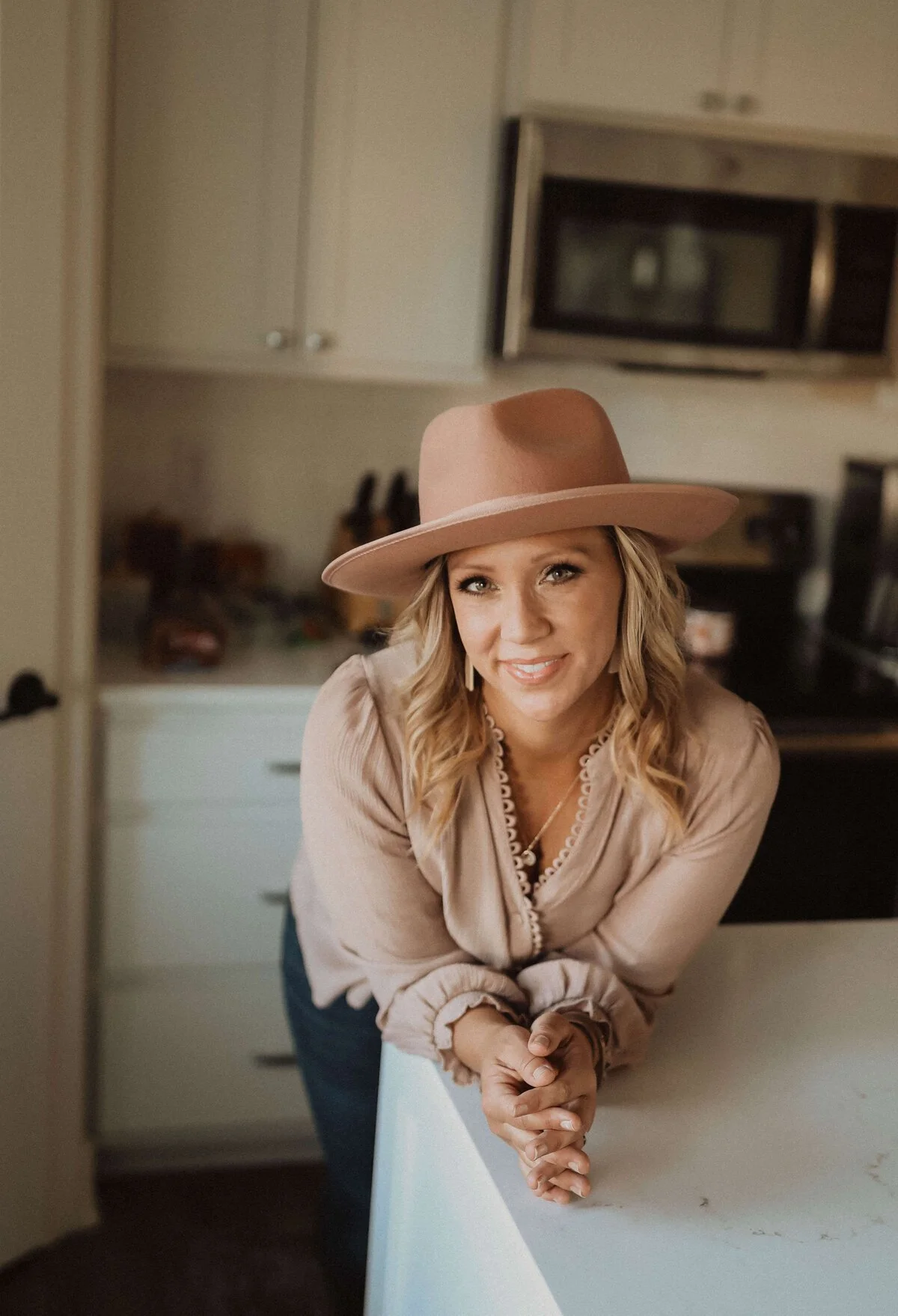 A woman with blonde curly hair wearing a beige wide-brim hat and beige blouse, smiling and leaning on a white kitchen counter in a home kitchen.