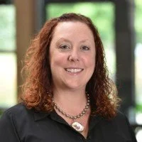 A man with curly dark hair, glasses, and a beard, looking to the side with a slight smile, wearing a dark green sweater