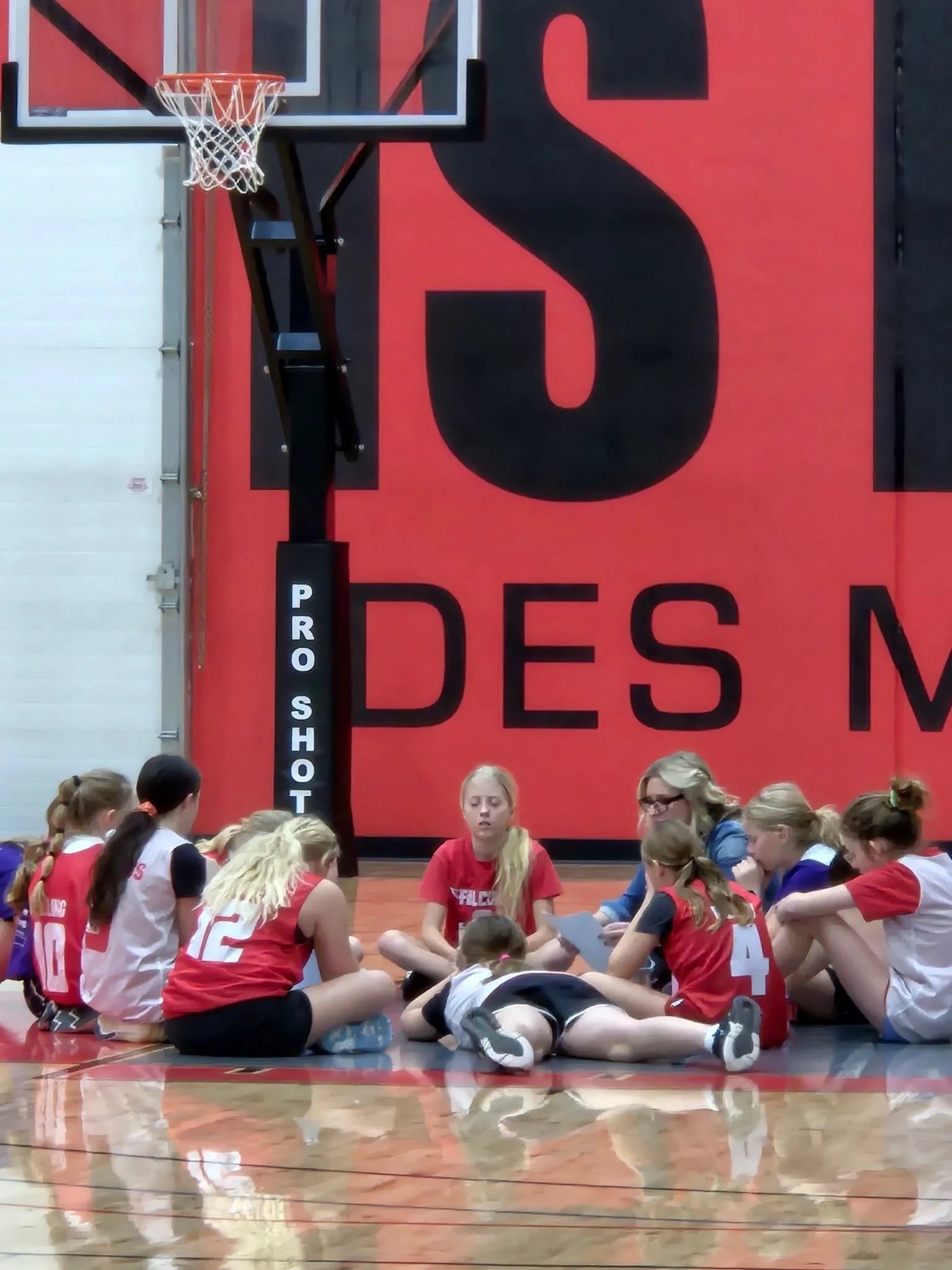 Group of young girls sitting in a circle on a gymnasium floor, with some reading papers and a coach or adult supervising, during a break in a volleyball game or practice session.