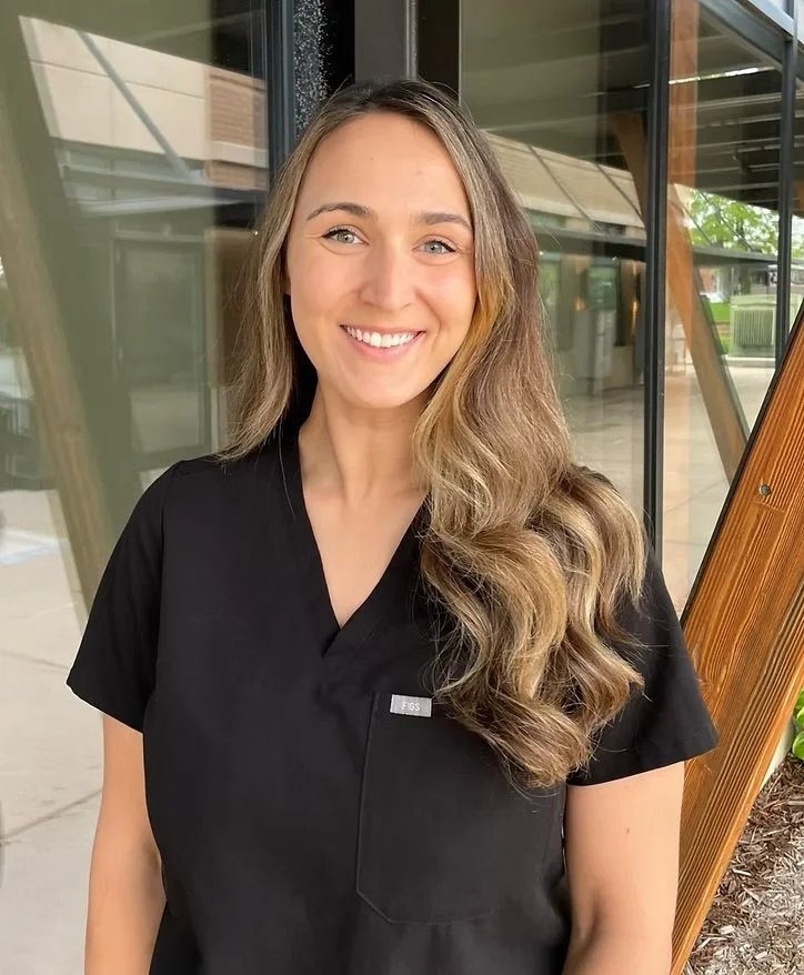 A woman wearing a black medical scrub top, smiling, with long, wavy light brown hair, standing outside near a glass building with wooden and green outdoor elements.