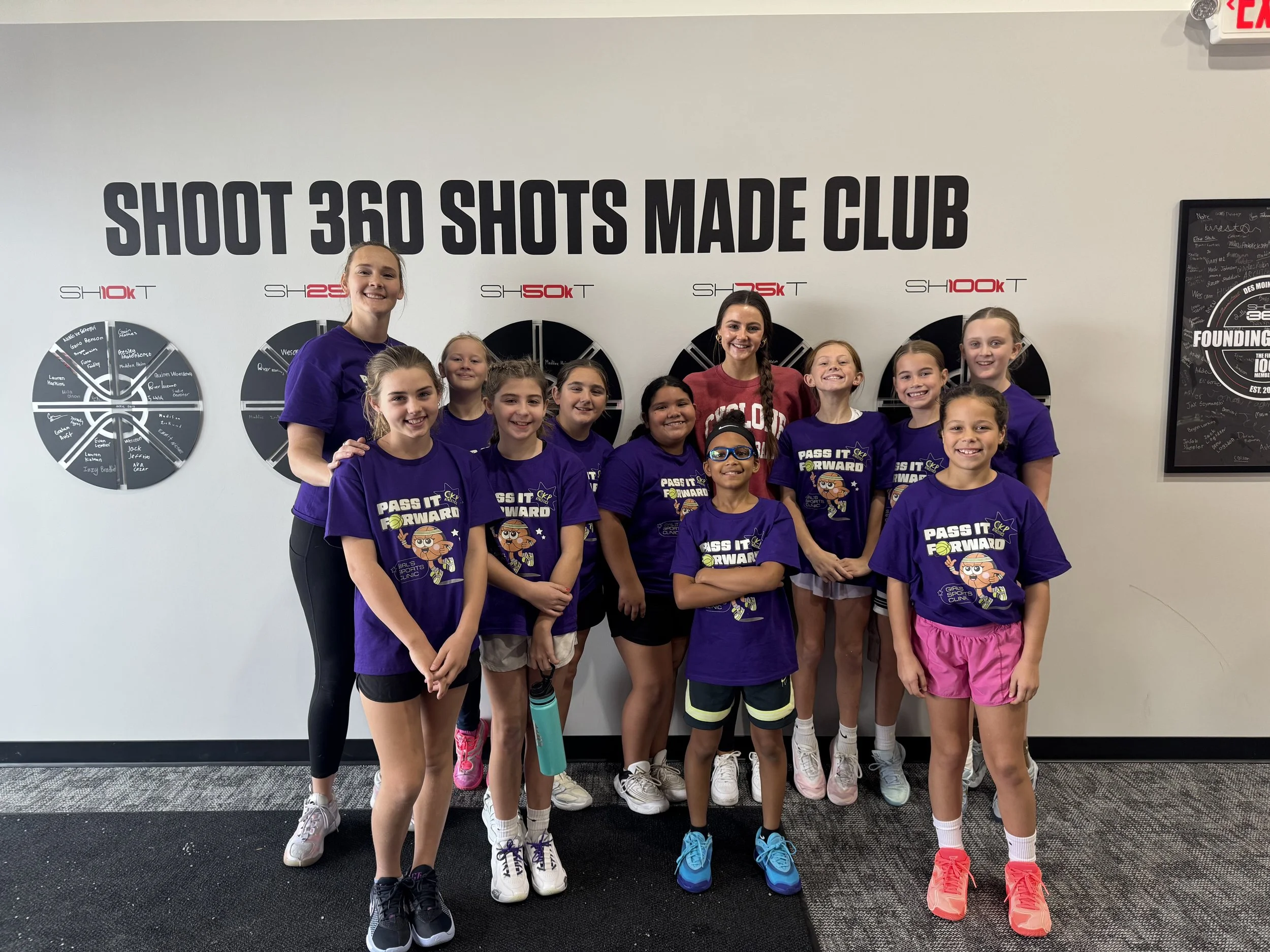 A group of young girls and two women standing in front of a wall with goal shots and motivational posters at a sports or fitness club. All are smiling, some wearing purple shirts with 'Pass It Forward' logo.