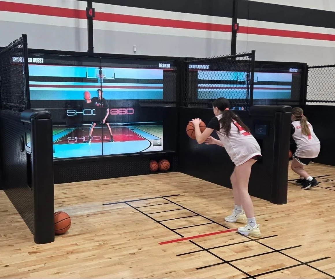 Two young girls in basketball uniforms playing a virtual basketball game at an indoor sports facility.