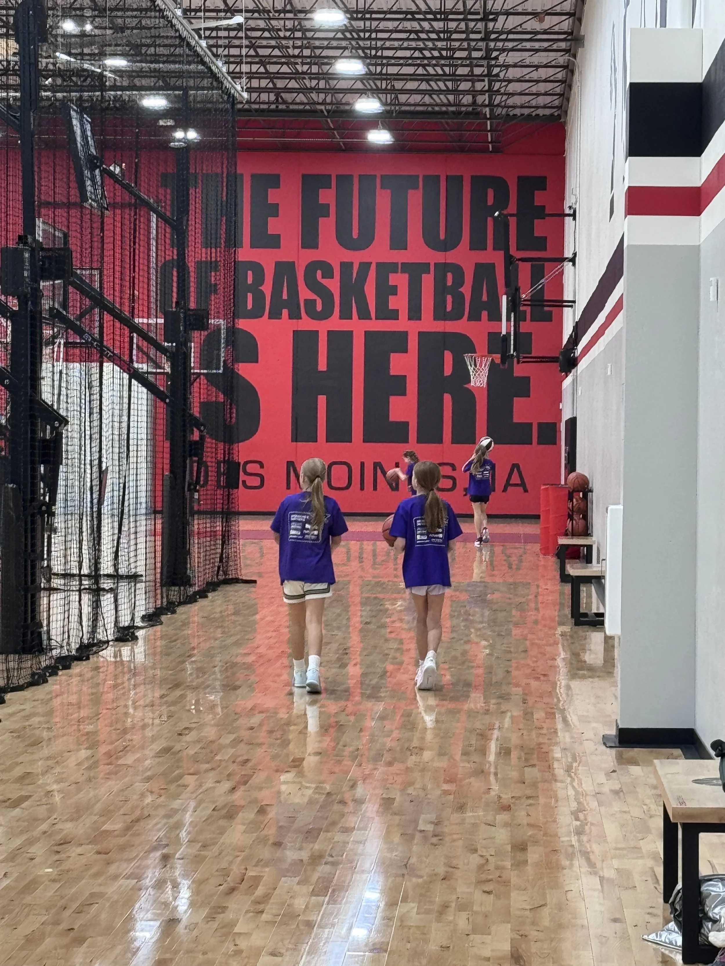 Children in purple T-shirts and shorts walking towards a basketball court with a wall that has large black letters saying 'THE FUTURE OF BASKETBALL IS HERE. COMING SOON TO A'.