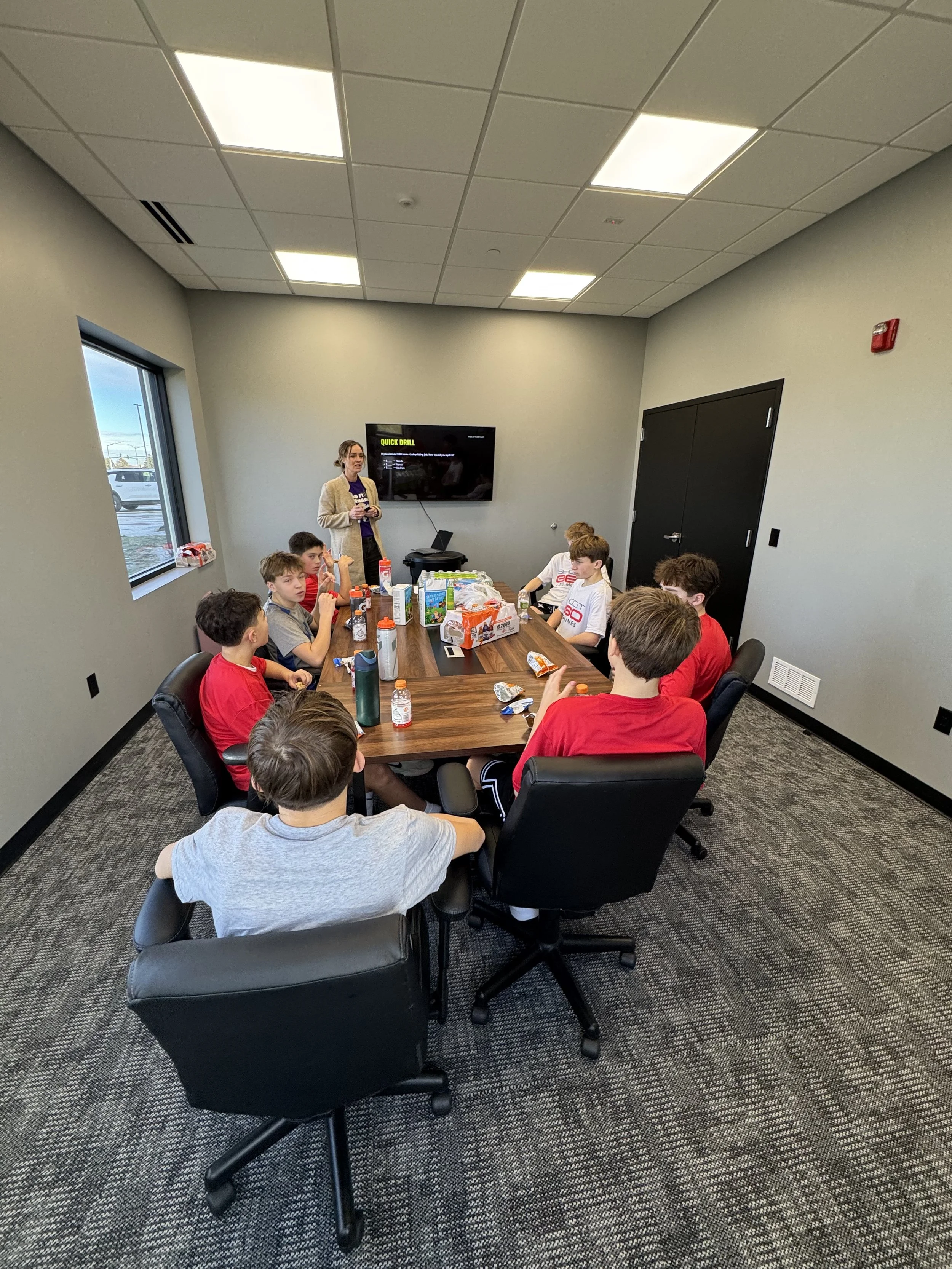 A group of kids sitting around a conference table with snacks, while a woman stands at the front giving a presentation in a meeting room.
