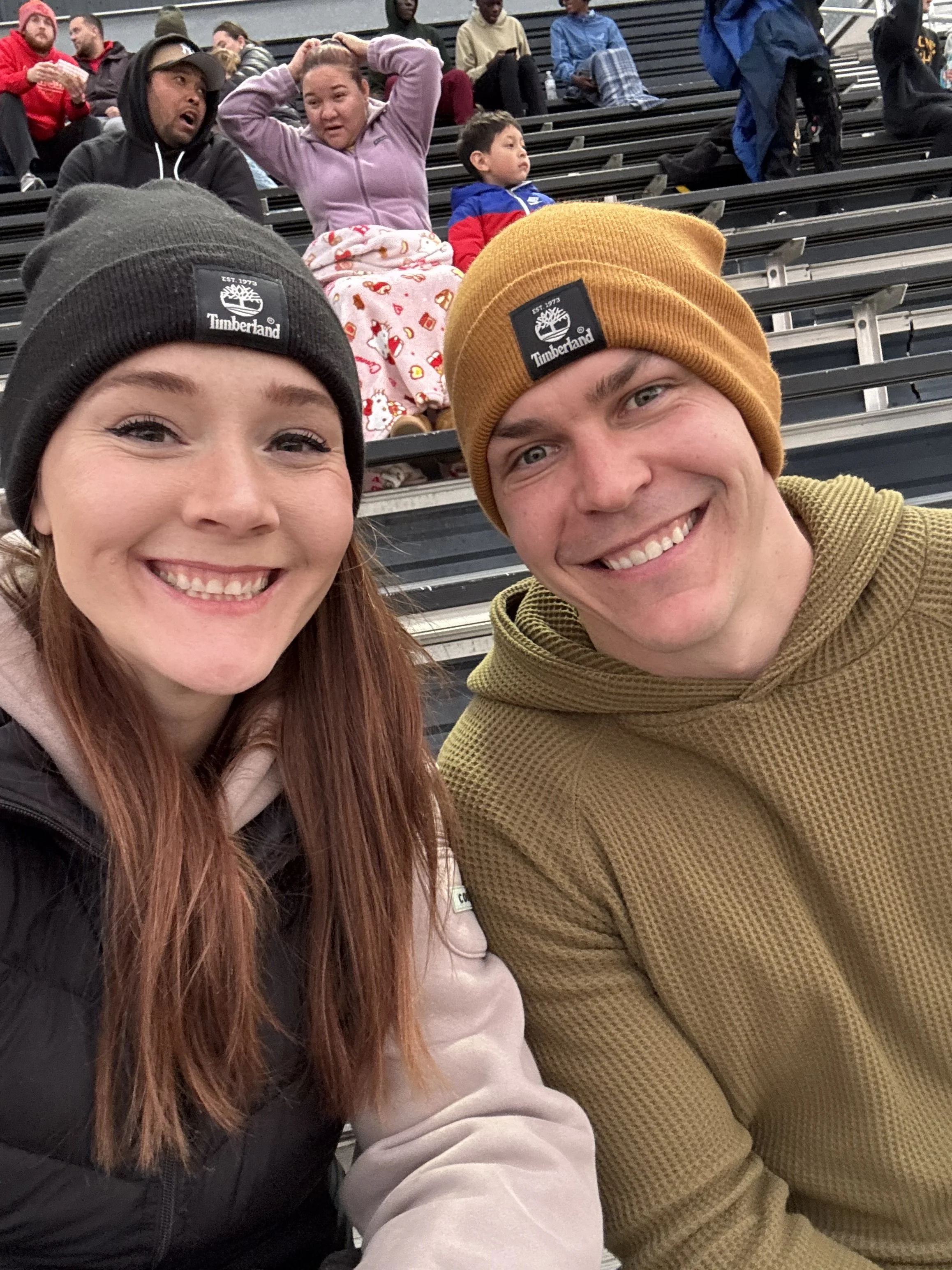 A smiling young woman and man taking a selfie in a stadium with people sitting on bleachers behind them.