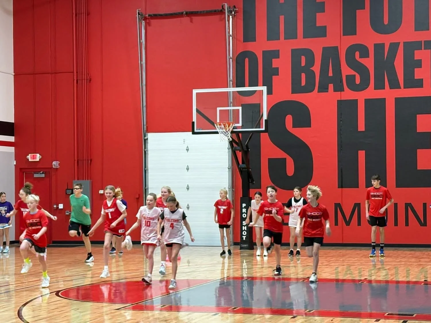 Group of young girls and boys playing basketball on an indoor court, with a red wall featuring large black text in the background.