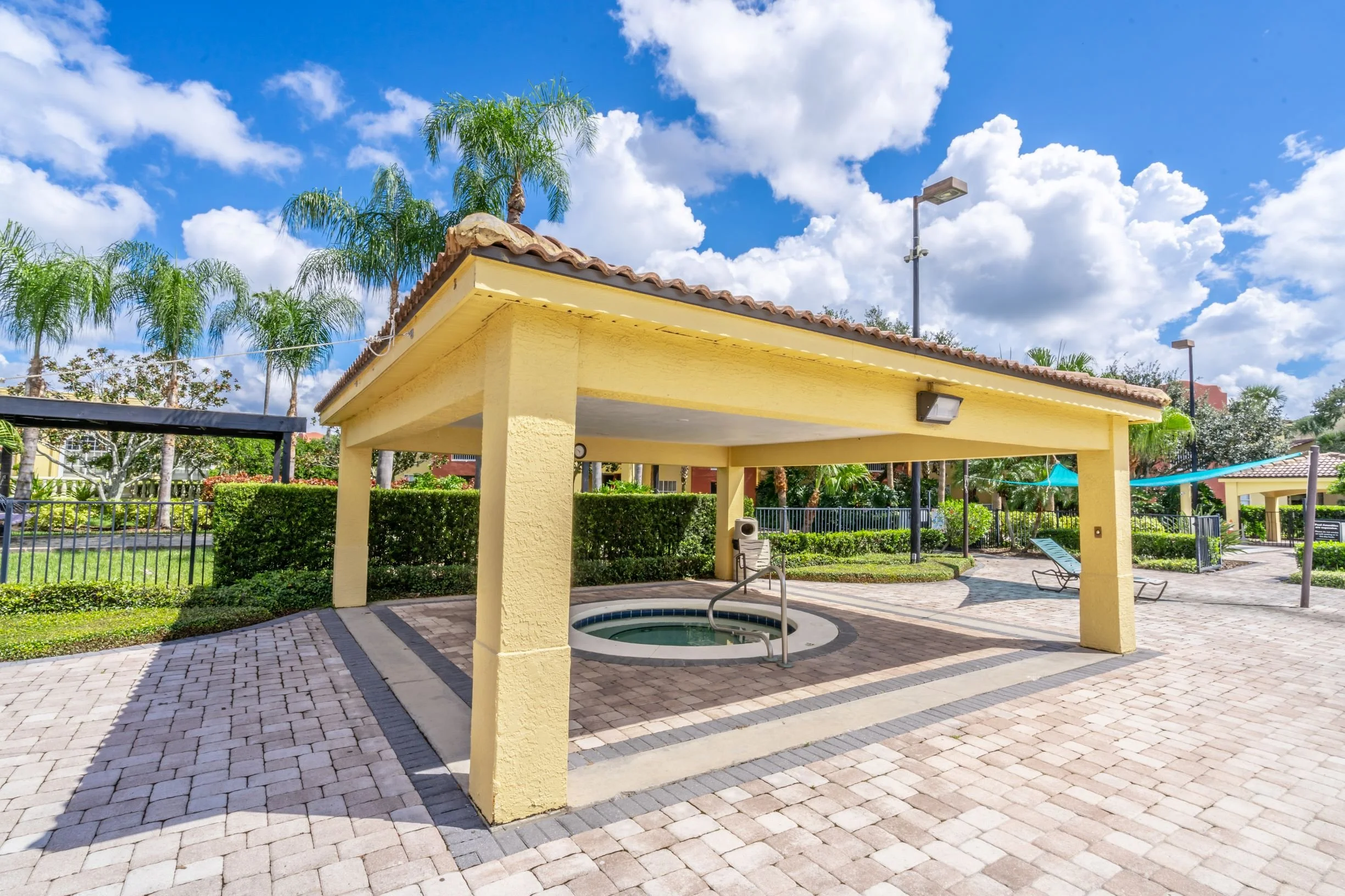 Outdoor hot tub beneath a yellow pavilion with landscaped greenery and palm trees in the background, blue sky with clouds, and paved walkway.