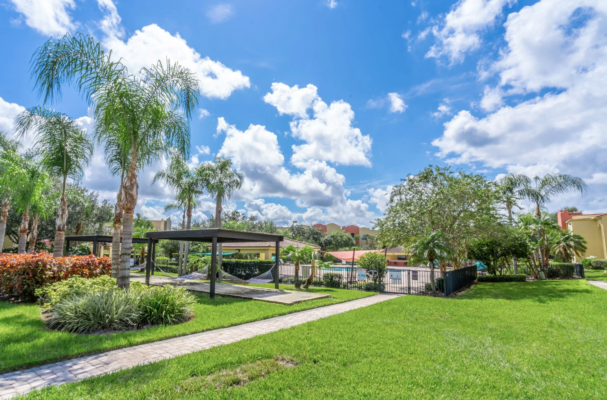 Sunny day at a park with green grass, palm trees, a shaded walkway, and a swimming pool area surrounded by a black fence.