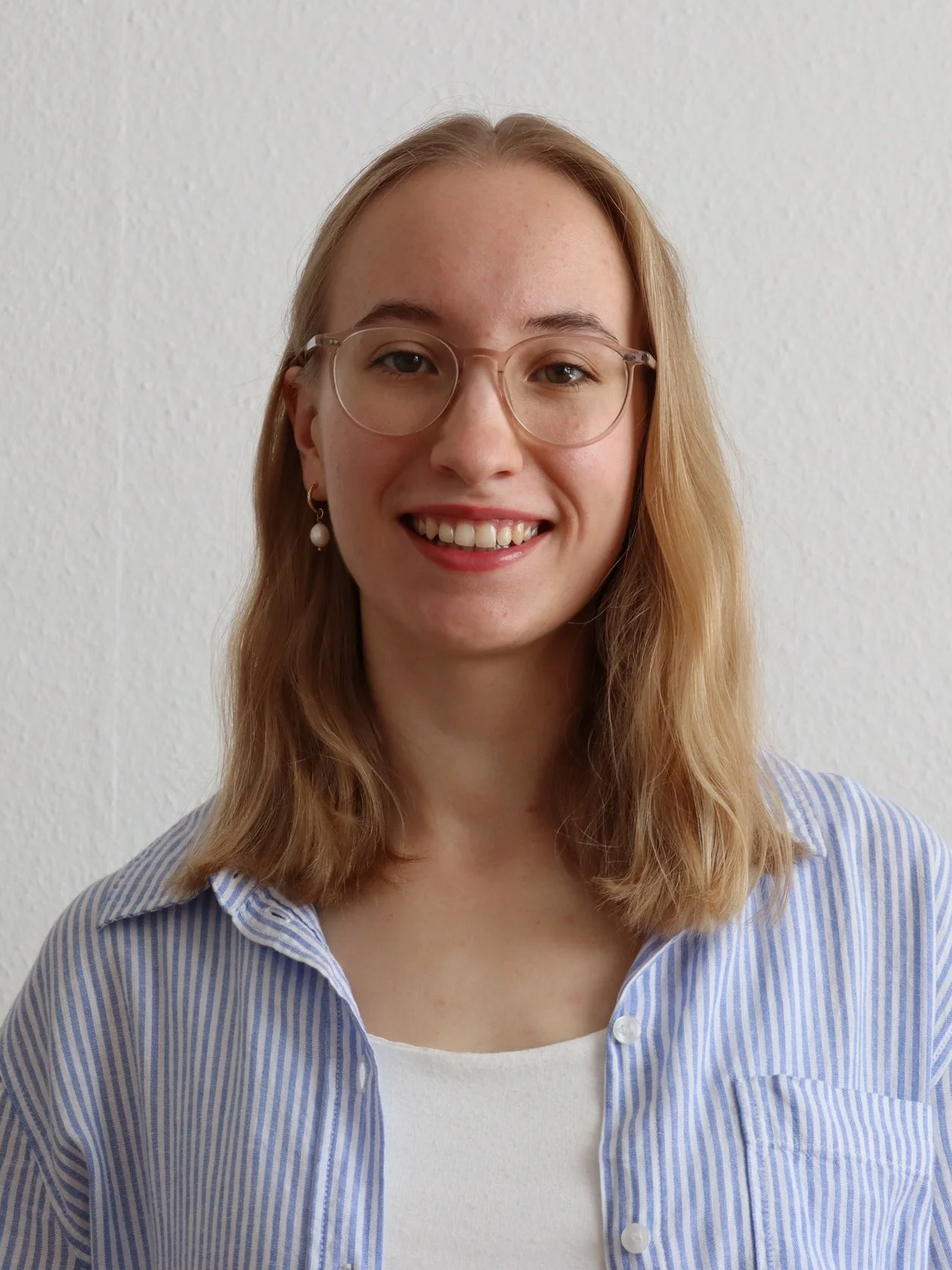 Porträt einer lächelnden jungen Frau mit Brille und Ohrschmuck vor weißer Wand, trägt ein blau-weiß gestreiftes Hemd.