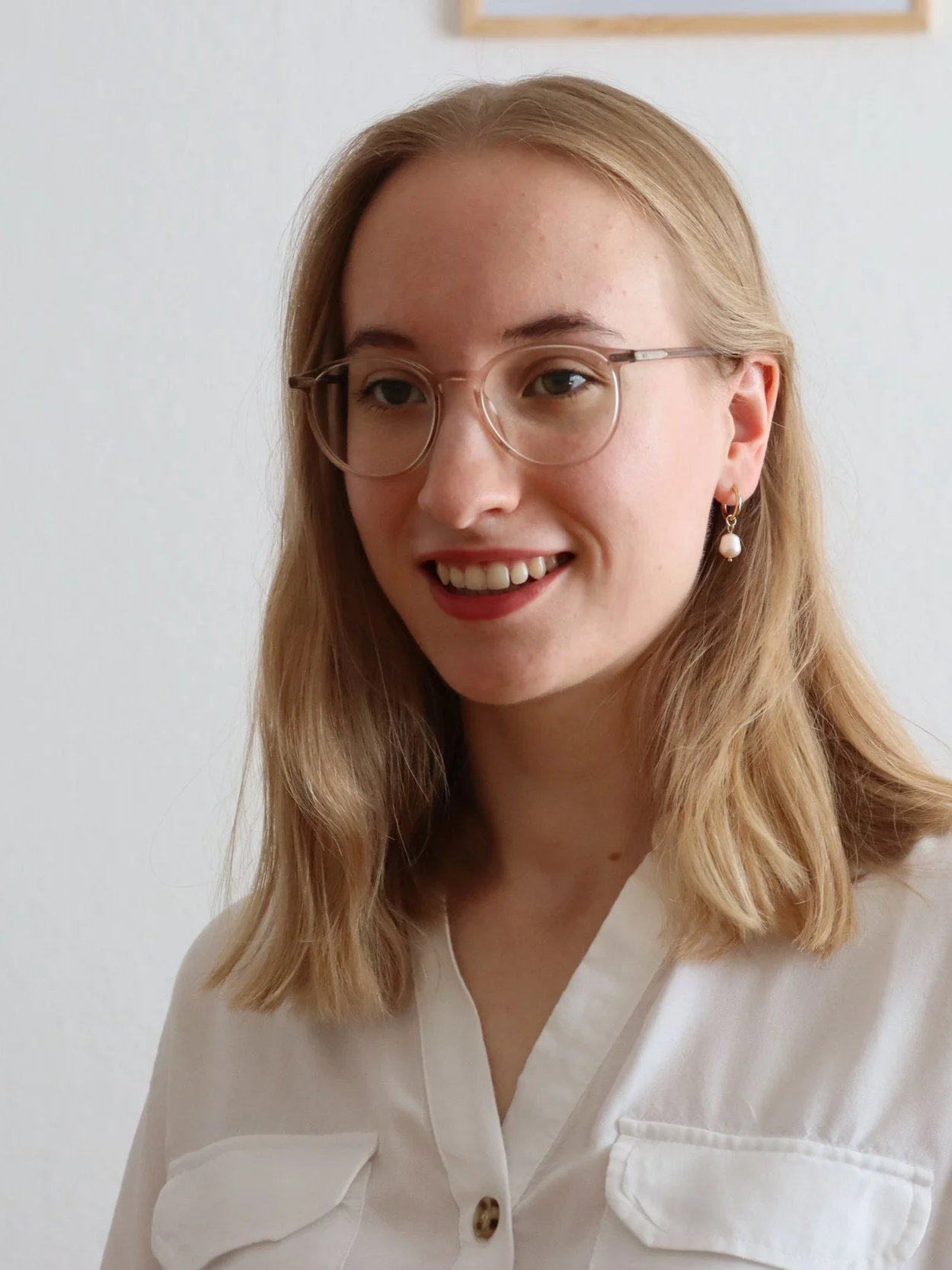 Junge Frau mit Brille und Perlenohrring, lächelnd, vor weißer Wand.