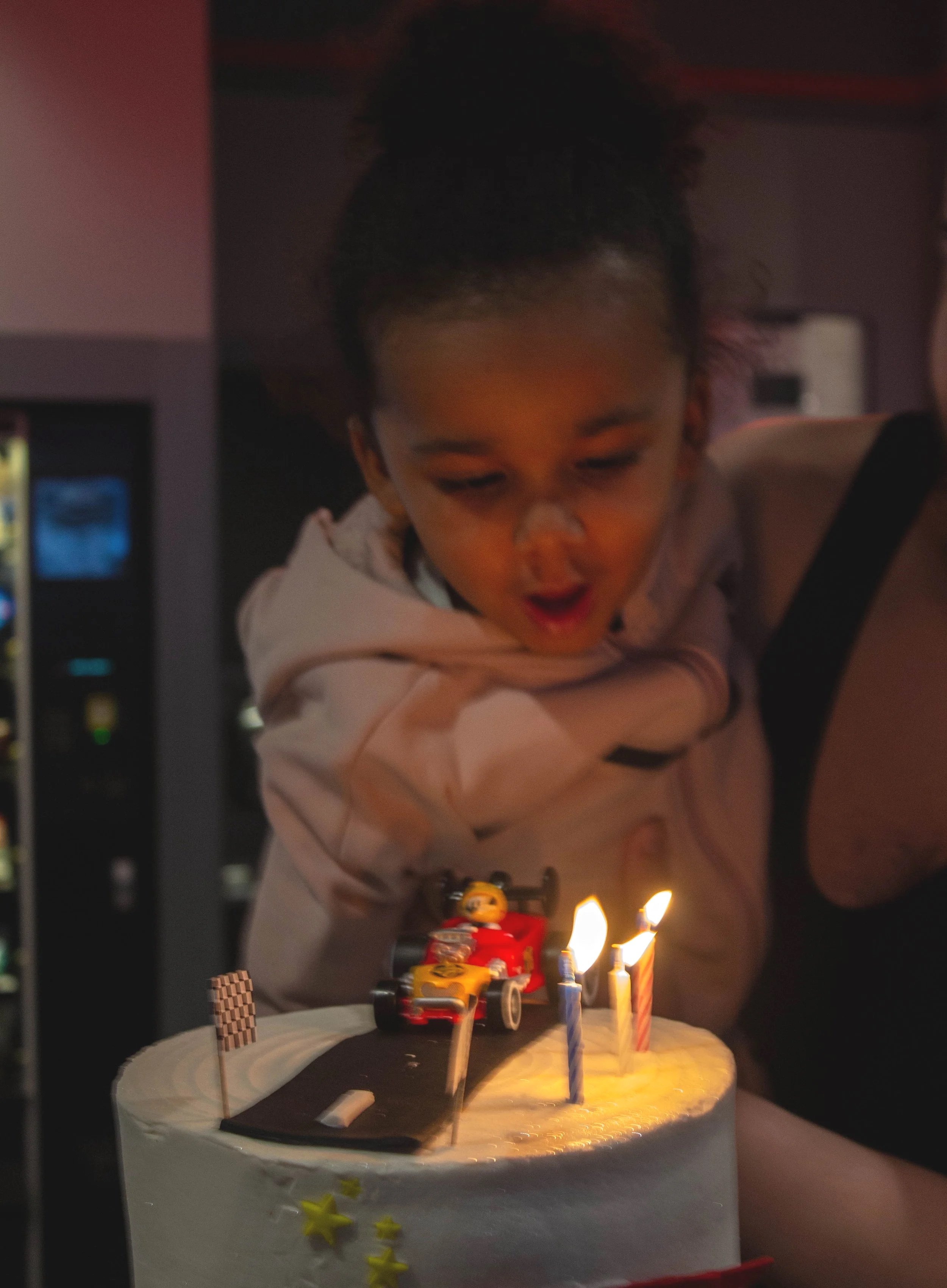 A child with curly hair is blowing out candles on a birthday cake decorated with a toy race car, small flag, and yellow stars.