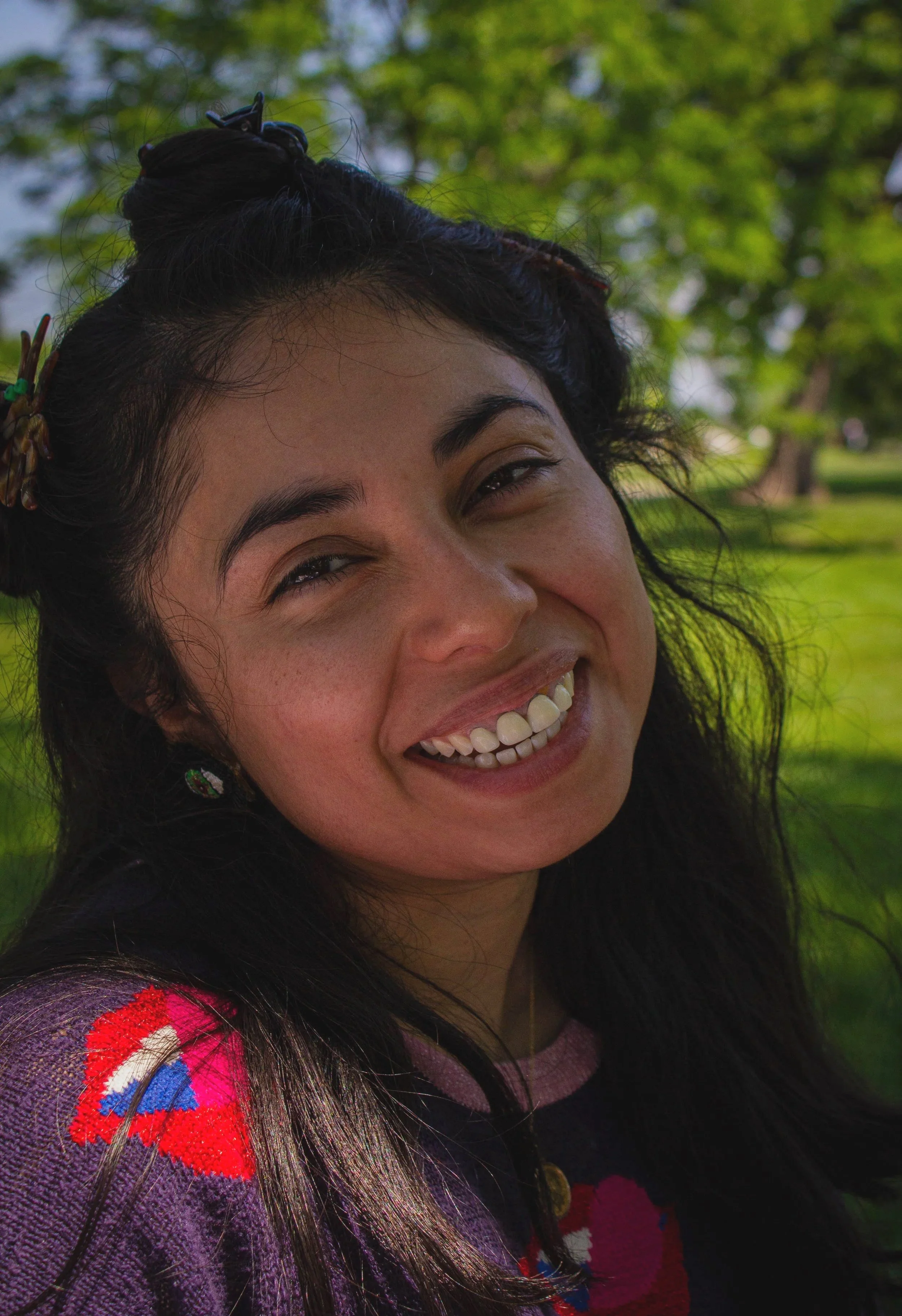 A person outdoors smiling, with dark wavy hair, wearing a multicolored sweater, in a park with green trees in the background.