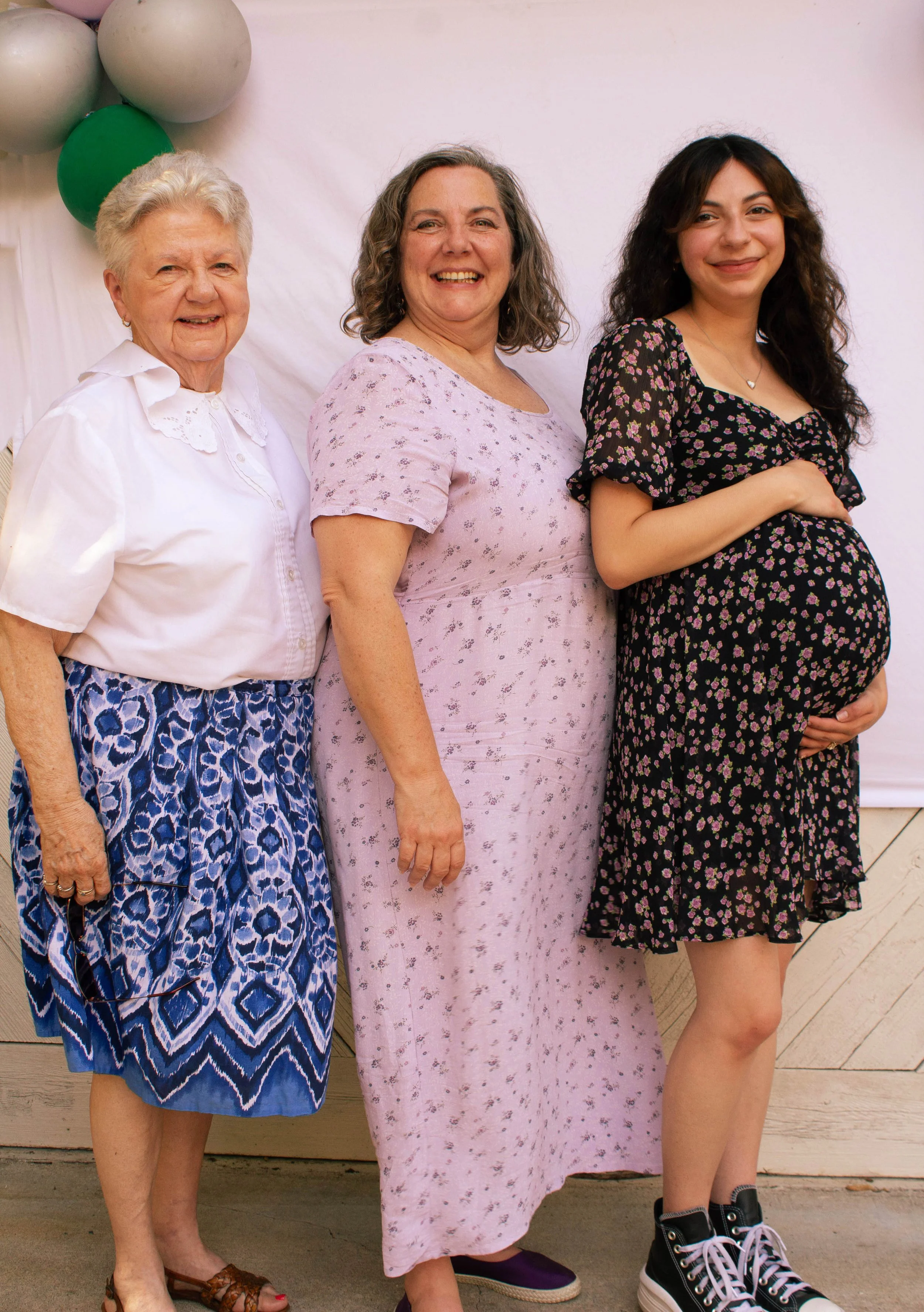 A young a pregnant woman on the right cradling her belly, standing side by side with her mother and grandmother. They stand in front of a white backdrop with balloons.