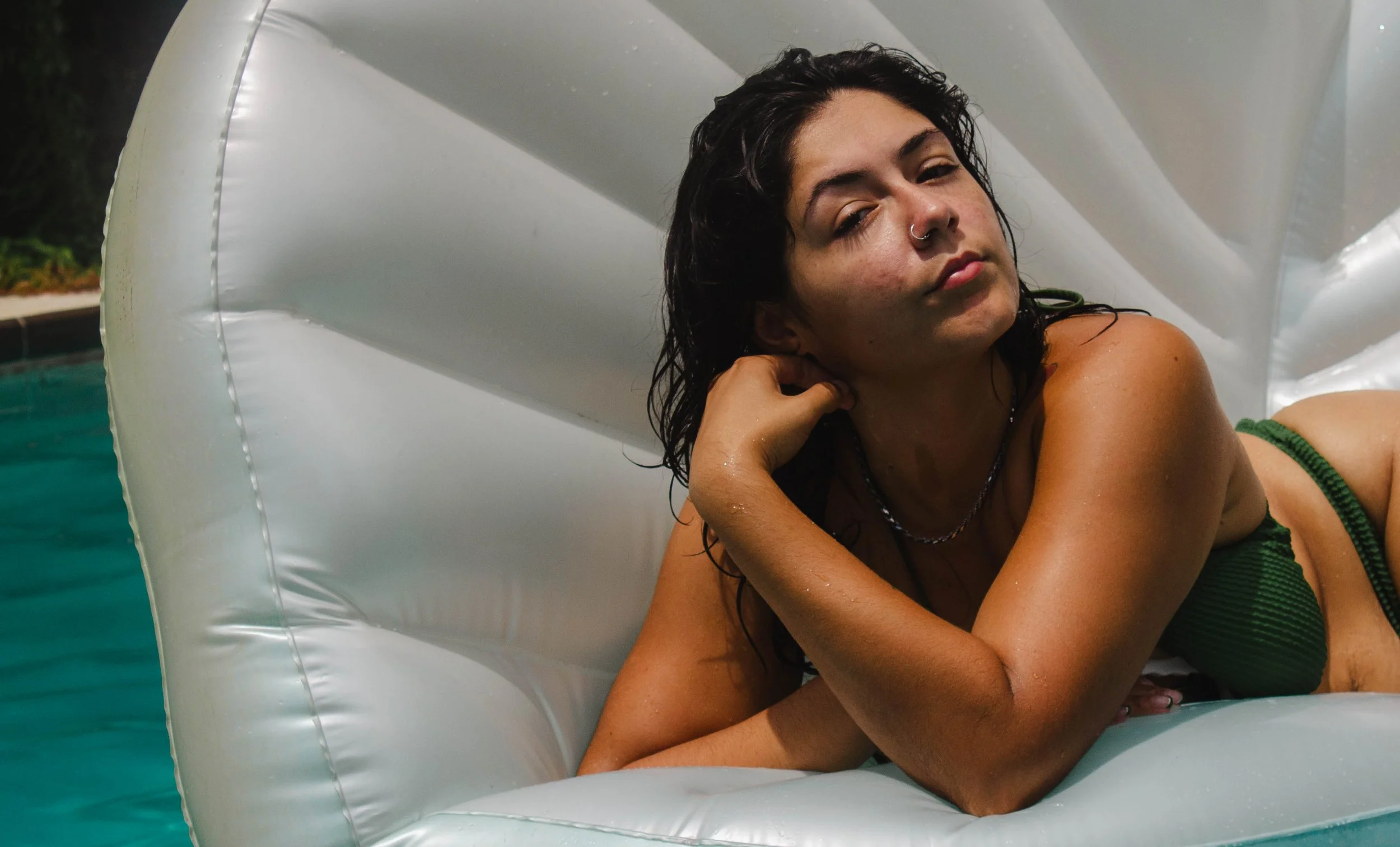 Young person with dark wet hair lying on an inflatable pool float in a swimming pool, resting their head on their hand with a relaxed and pensive expression.