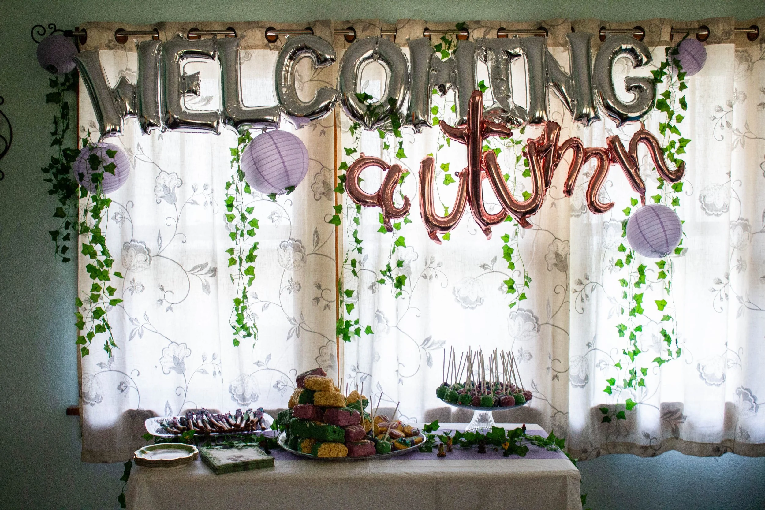 Picture of a pastry display table for a baby shower. The pastry table sits in front of large windows. Over the windows, nylon balloons spell out "welcoming Autumn"