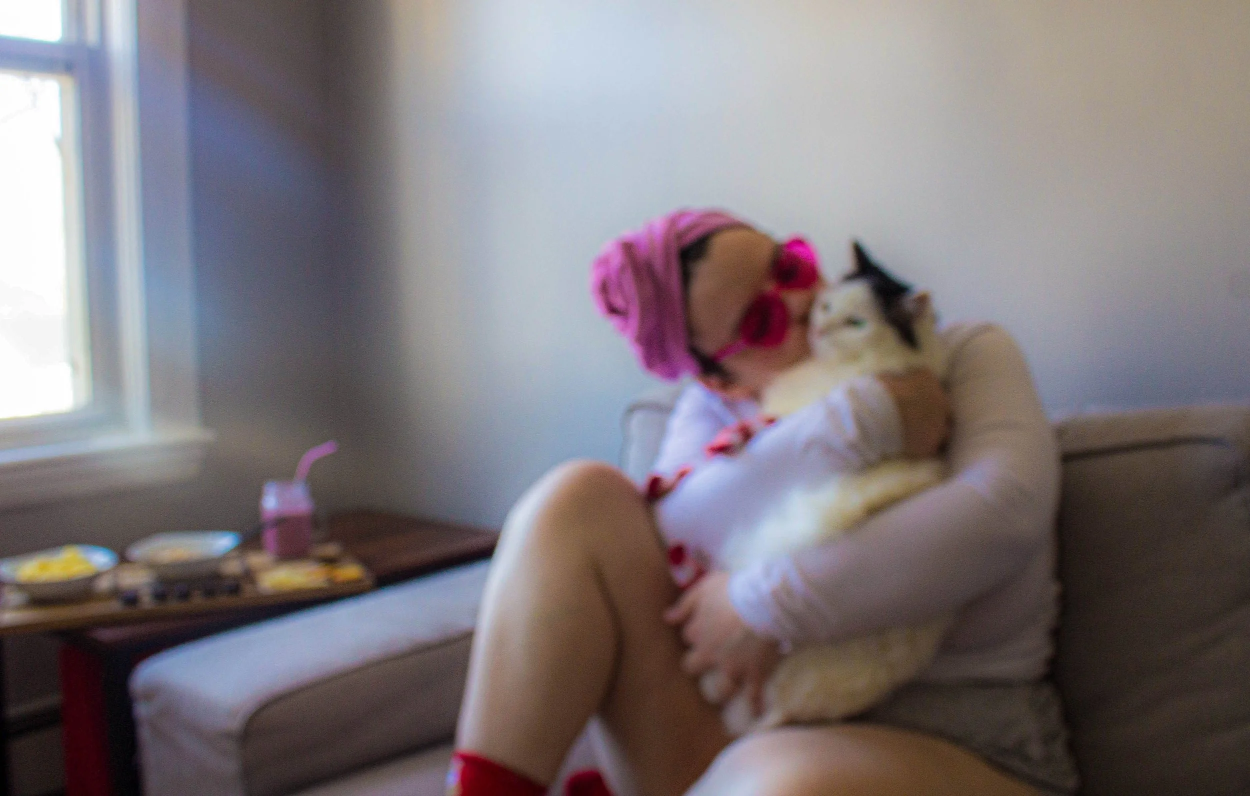 A person is sitting criss-crossed on a couch with one leg propped up. They wear a pink hair towel, pink glasses and red socks. They are holding their white cat and giving the cat a kiss. In the background, a tray of food is displayed.