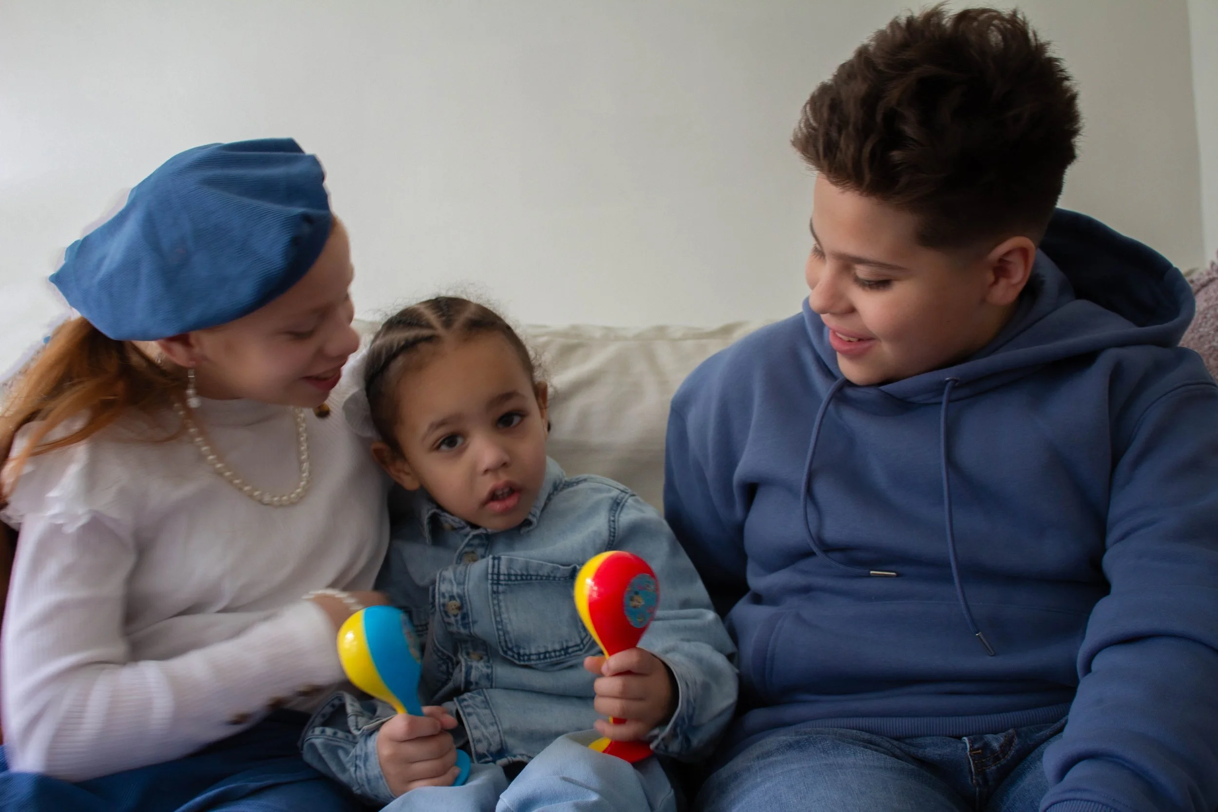 Three children sitting on a couch, engaging in a playful activity with maracas, smiling and looking at each other, in a cozy indoor setting.