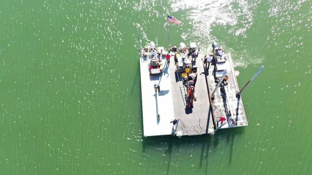 An aerial view of a flat boat with people on board, floating on green water, with an American flag on the boat.