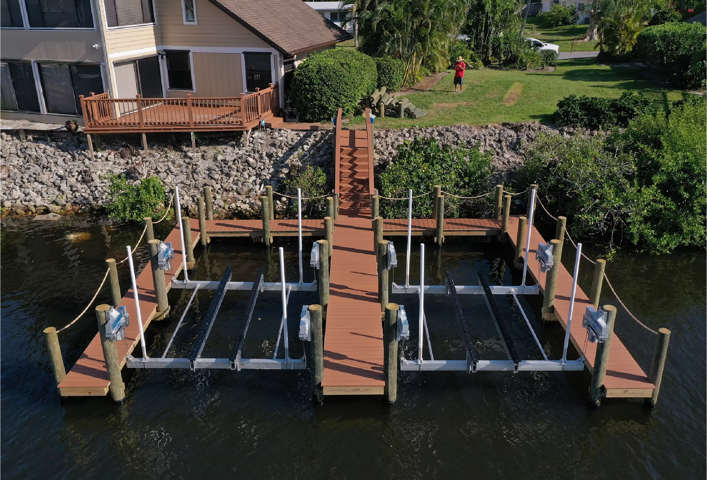 A dock structure with multiple boat slips and cleats, situated on calm water, leading to a residential yard with a deck, bushes, and a person taking a photograph.