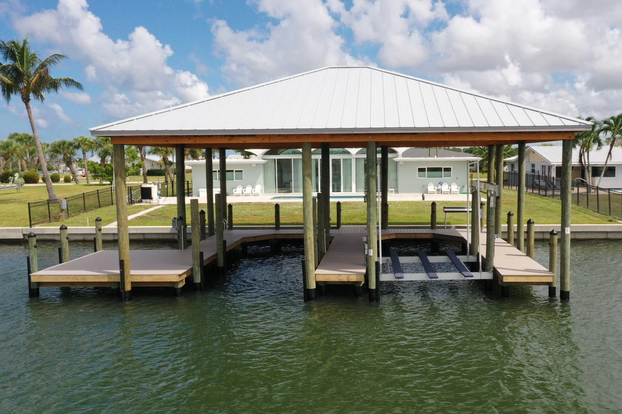 A wooden dock with a covered roof extending over water, leading to a backyard with a house, lawn, and palm trees under a partly cloudy sky.