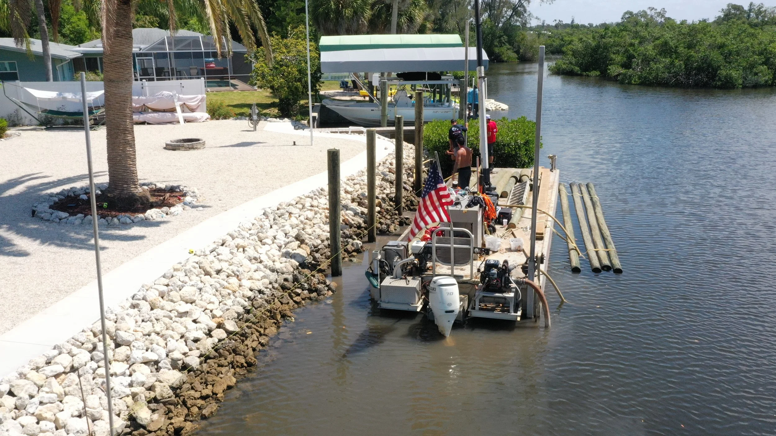 A small dock with bamboo poles extends into a body of water next to a landscaped yard with a palm tree and a fire pit. People are working on a boat at the dock, which is equipped with various gear and an American flag.