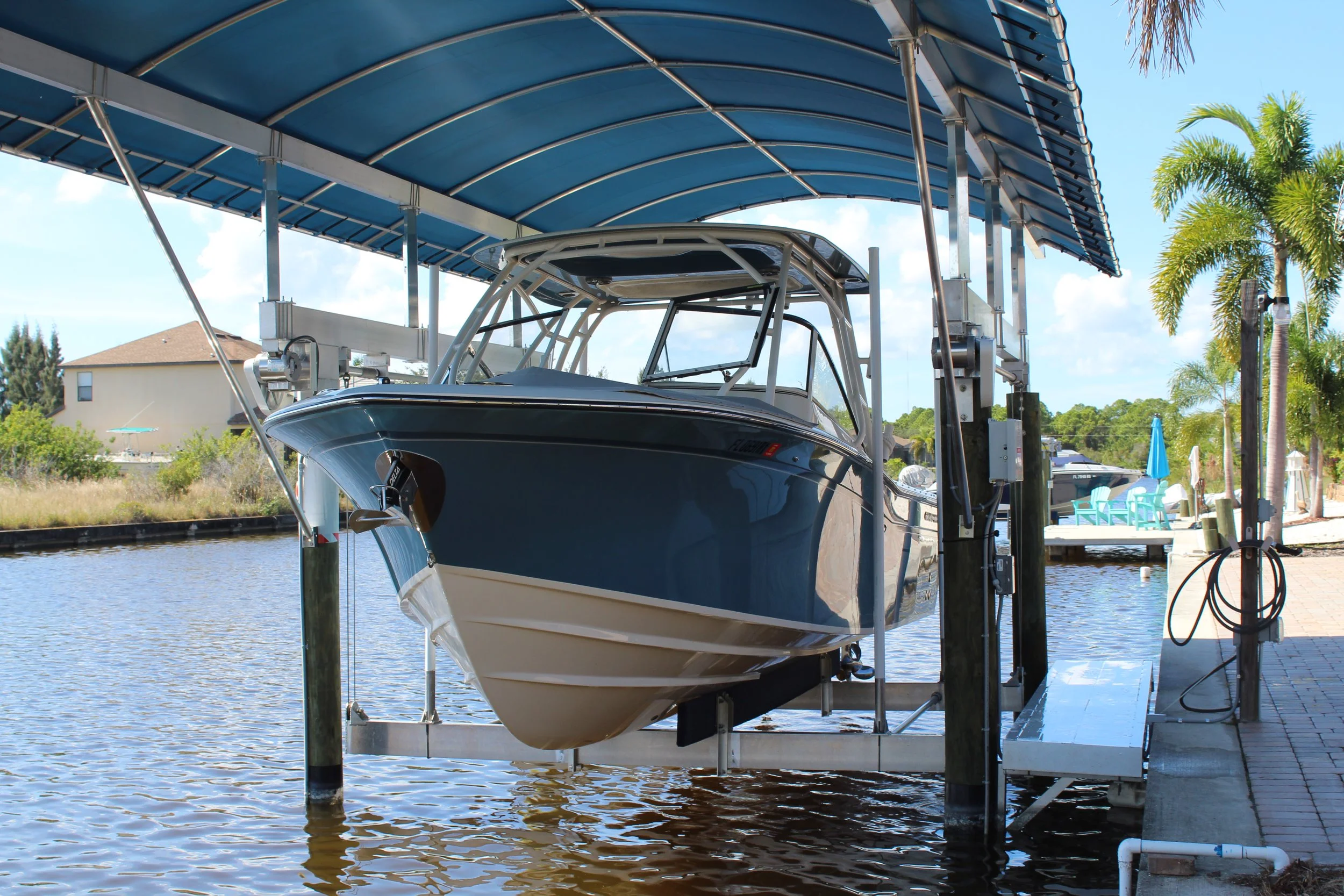 A boat stored on a lift under a roof over a water canal, with trees and neighboring houses in the background.