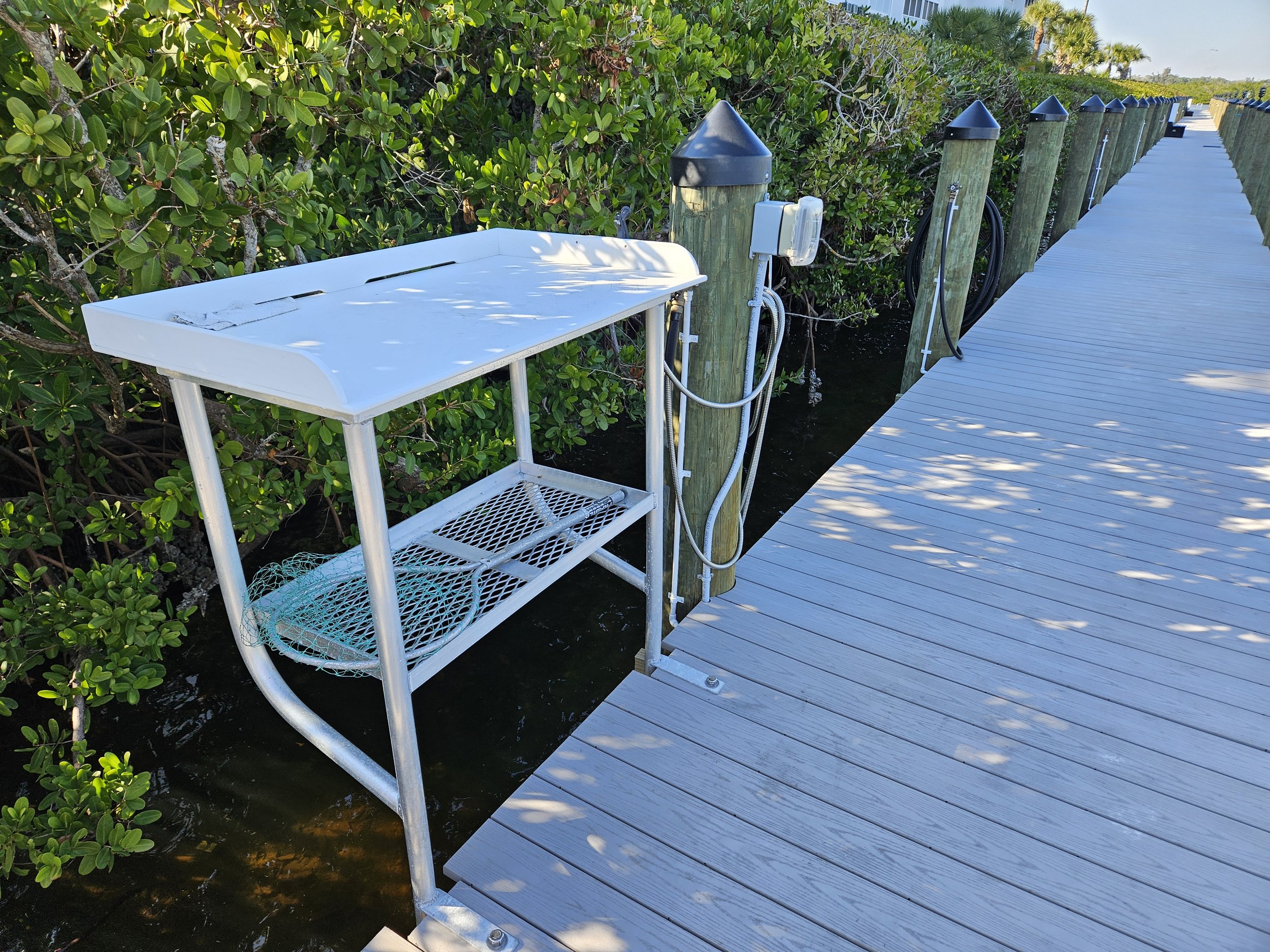 A white boat dock with a metal fish cleaning table and an electrical outlet, surrounded by green bushes, with water underneath the dock and poles along the pier.