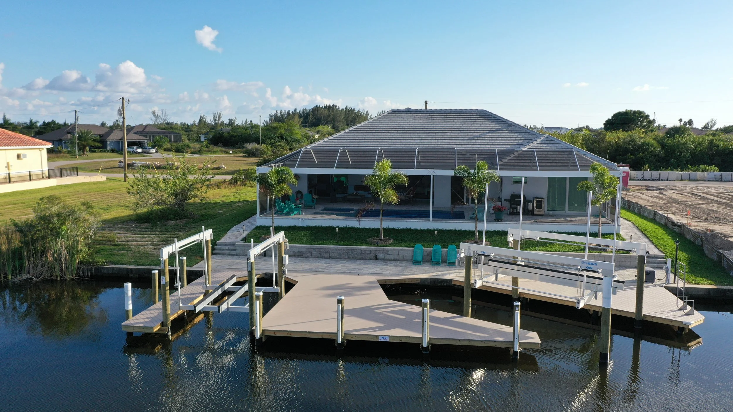 A house with a screened porch overlooking a canal with a dock and boat lifts, surrounded by green lawn and trees, under a bright blue sky with some clouds.