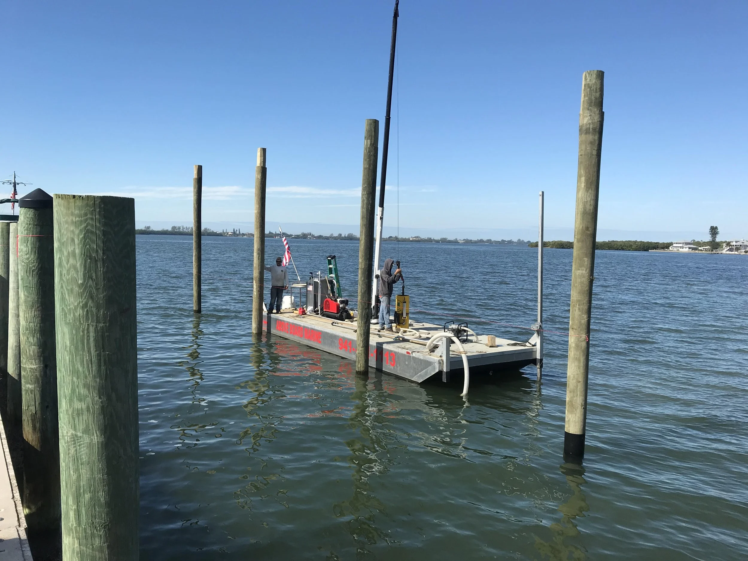 A floating dock at a body of water with two people preparing equipment, surrounded by wooden pilings. The scene shows a clear blue sky.