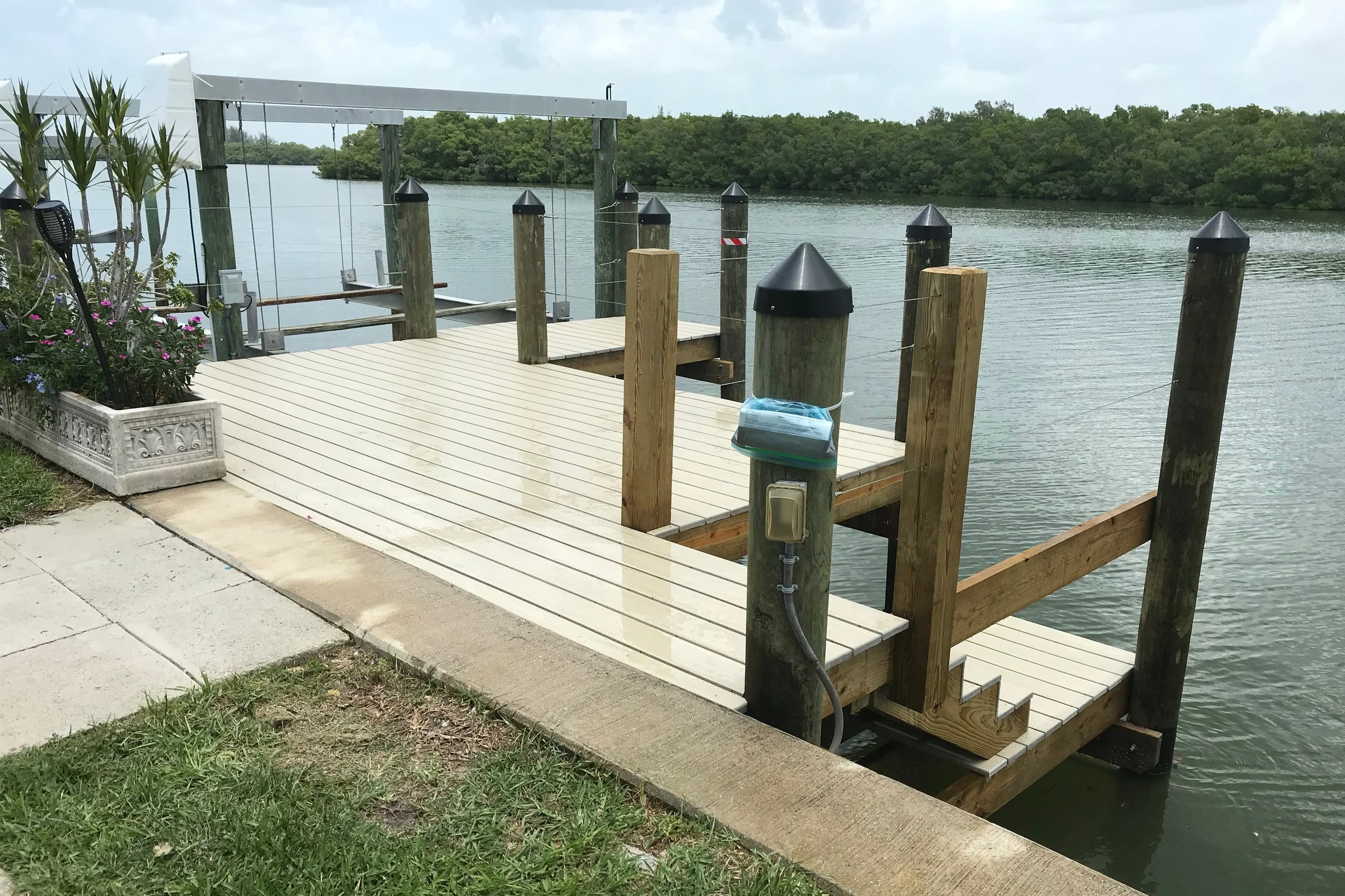 A newly built wooden dock extending over a body of water with boat cleats, a mailbox, and a power outlet. Surrounding vegetation and a cloudy sky are also visible.