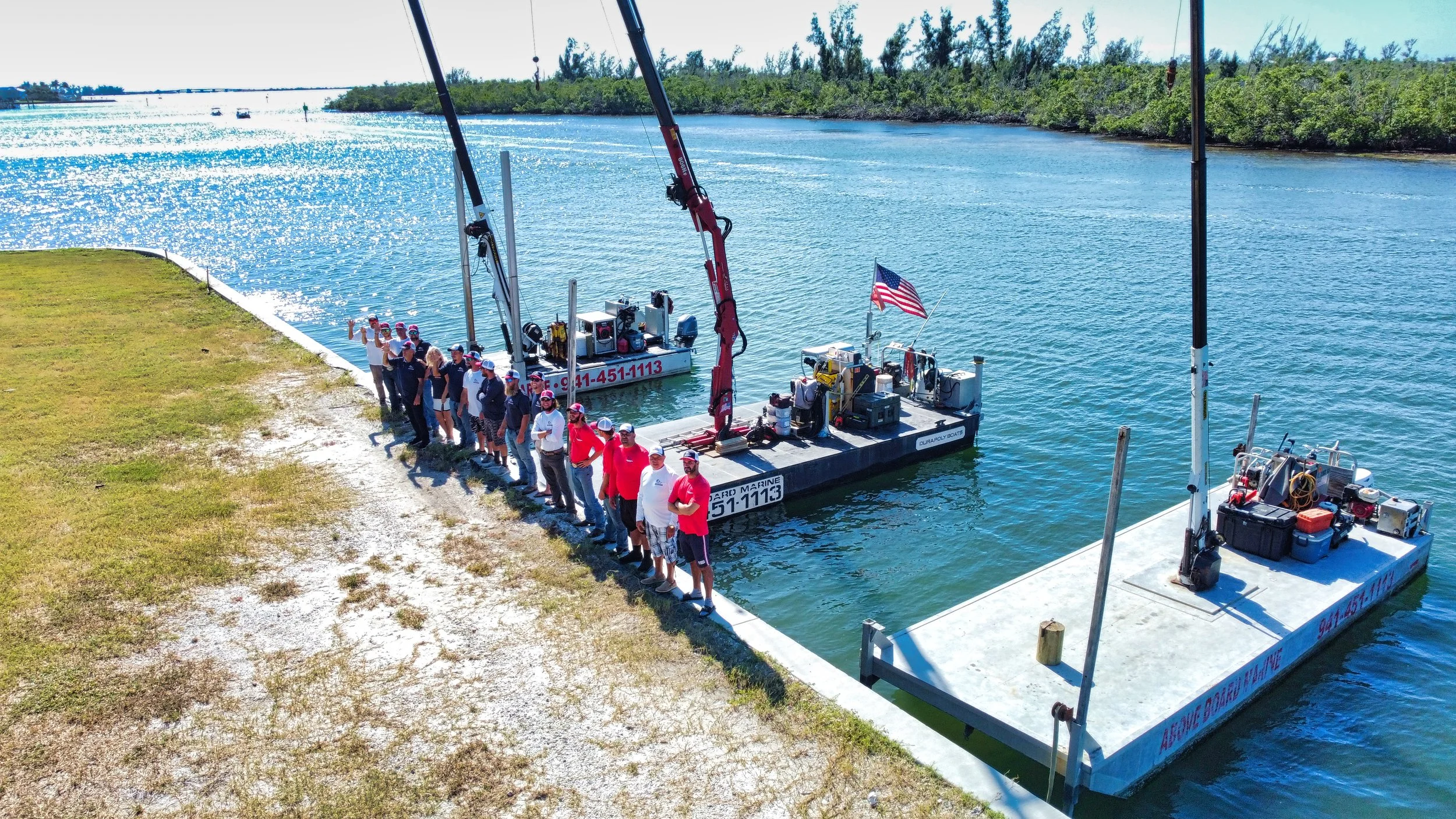 Group of people standing on a shoreline next to two large flat boats in the water, equipped with cranes and machinery, with a US flag flying on one boat. The scene is bright and sunny, with water in the background and boats visible on the water.