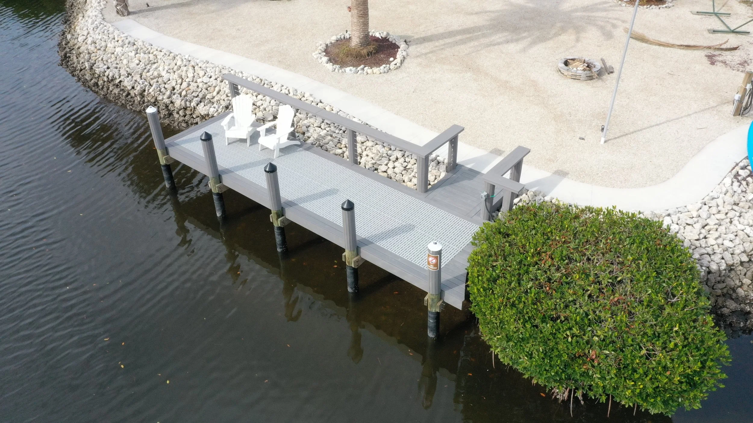 A small dock with white chairs extends over a body of water beside a landscaped area with a tree and bush.