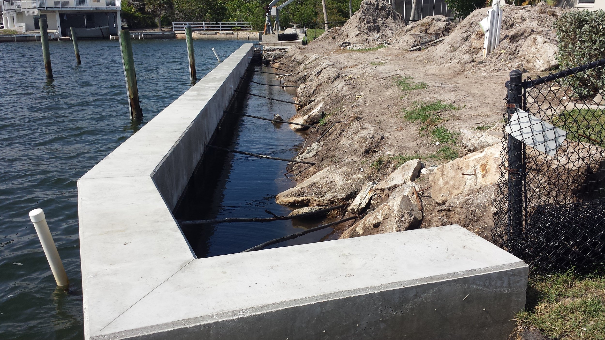 Construction site with a concrete seawall and a partially filled water channel along a shoreline, rocks and dirt on the land side, a chain-link fence on the right, and a utility pipe in the water on the left.