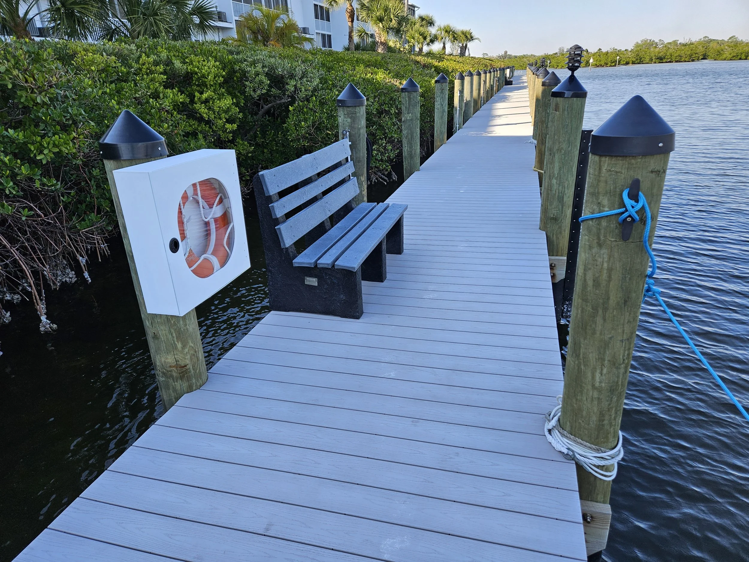 A wooden dock extends over the water with a bench and a white life ring storage box attached to a post. The dock is secured by ropes tied to the posts, and lush green bushes and palm trees line the waterfront.