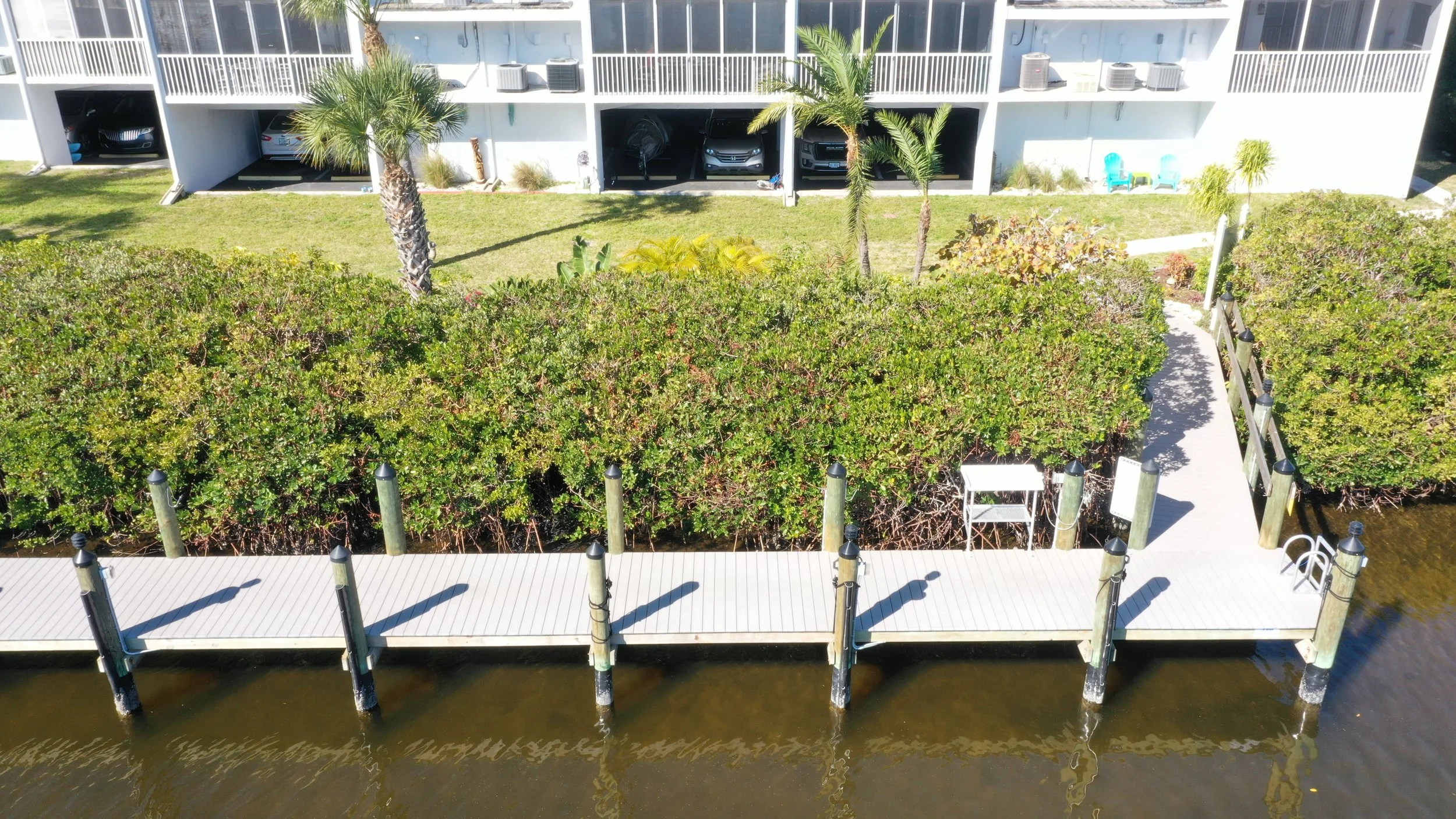 Aerial view of a waterfront residential area with a wooden dock extending into the water, surrounded by green shrubs and palm trees, with a white multi-story building with parking garages in the background.