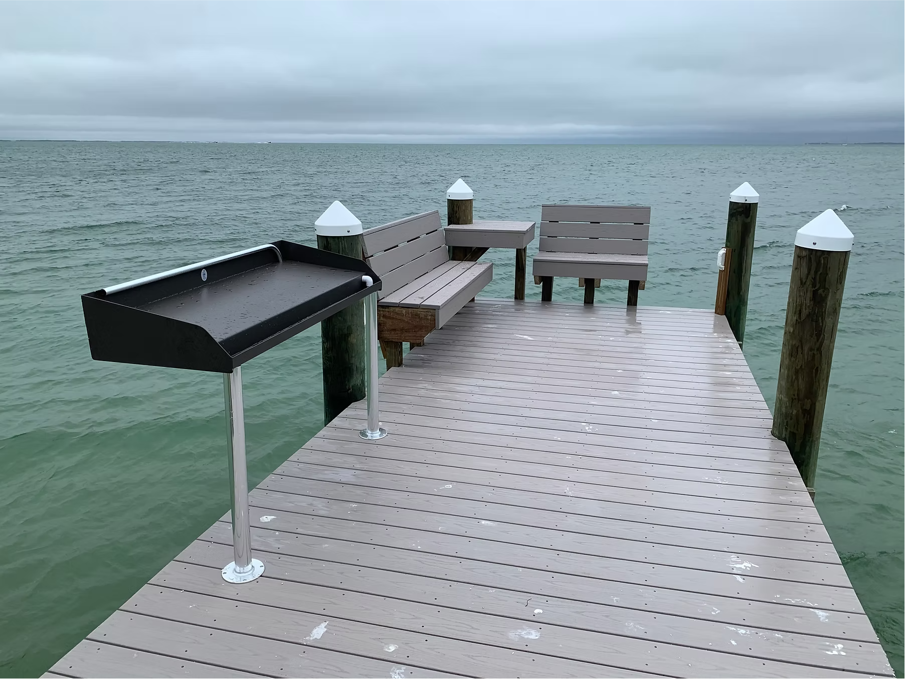 A wooden pier extends over calm water with a view of cloudy sky. The pier has benches, a trash receptacle, and white-topped posts.