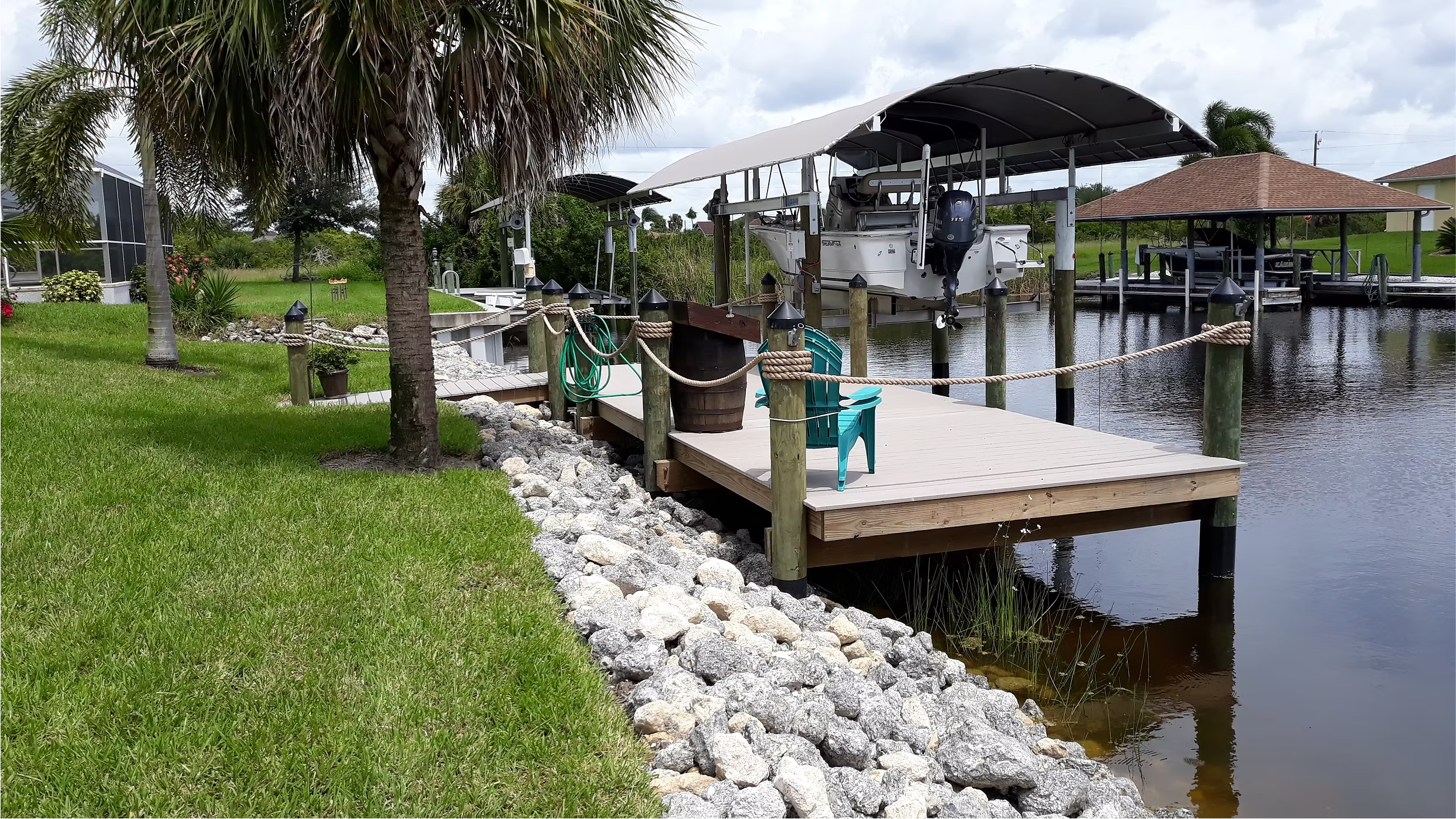 A wooden dock with ropes and posts extending into a calm water canal, featuring a covered boat lift with a boat and engine, surrounded by green grass, palm trees, and residential houses.