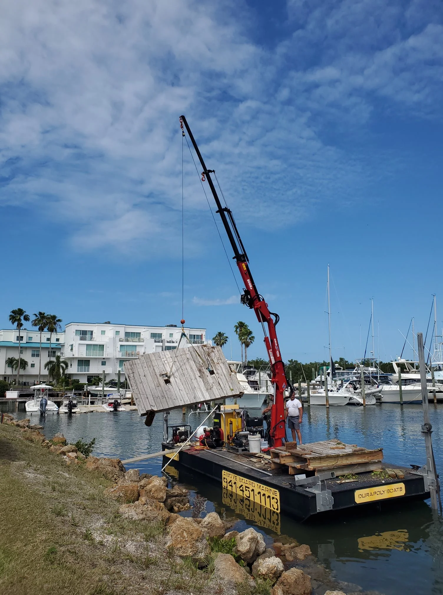 A small boat with a crane lifting a wooden panel near a marina with boats and sailboats docked, and a white building with palm trees in the background.