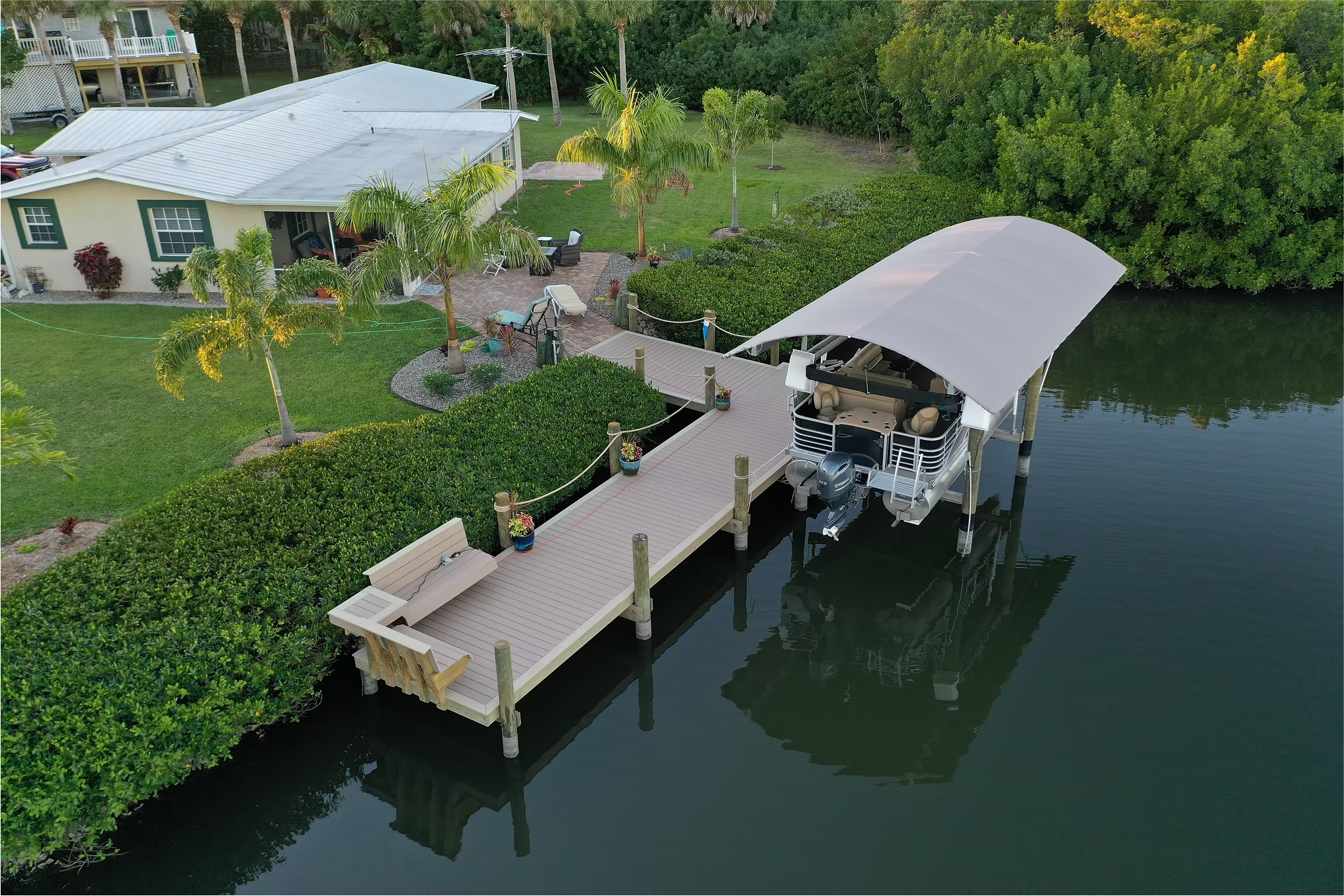 A dock with a boat covered by a white canopy, extending from a backyard with palm trees, patio furniture, and a house with a white roof, along a calm waterway surrounded by lush greenery.