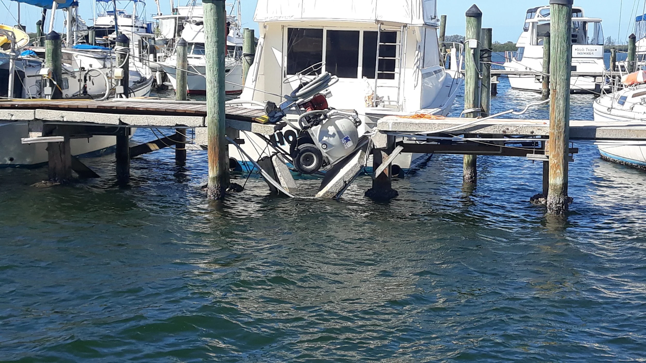 A boat is partially submerged in water at a marina, caught between a dock and the sea, with visible damage and a motor on the front. Multiple other boats are docked nearby.