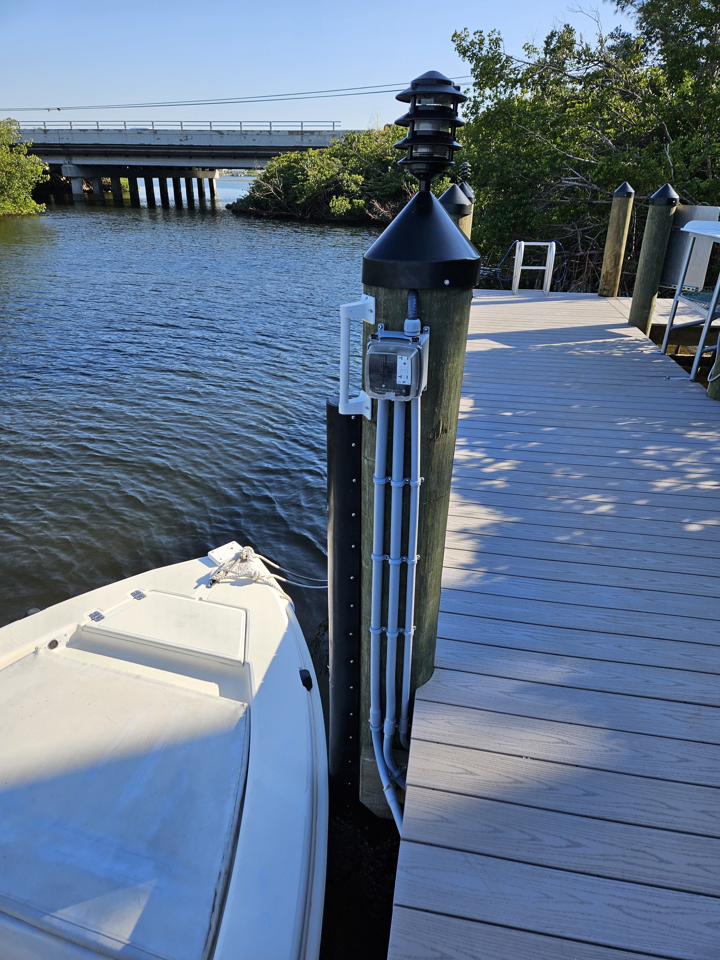 A boat dock shows a utility pole with electrical and possibly data lines, next to a boat tied to the dock, with water and trees in the background.